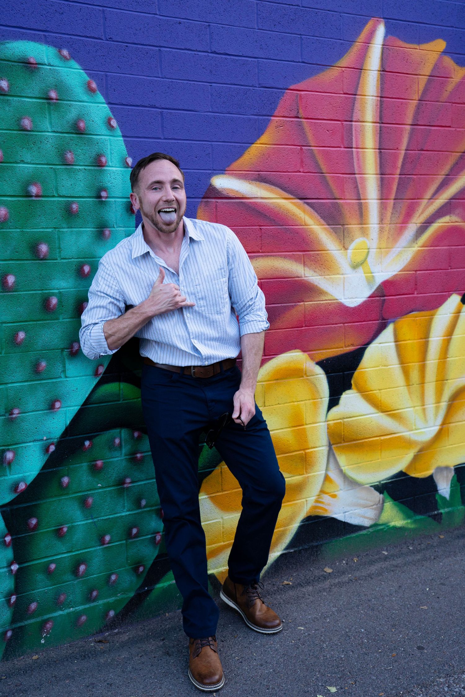 Man laughing in striped shirt leaning against colorful cactus and floral mural on brick wall.