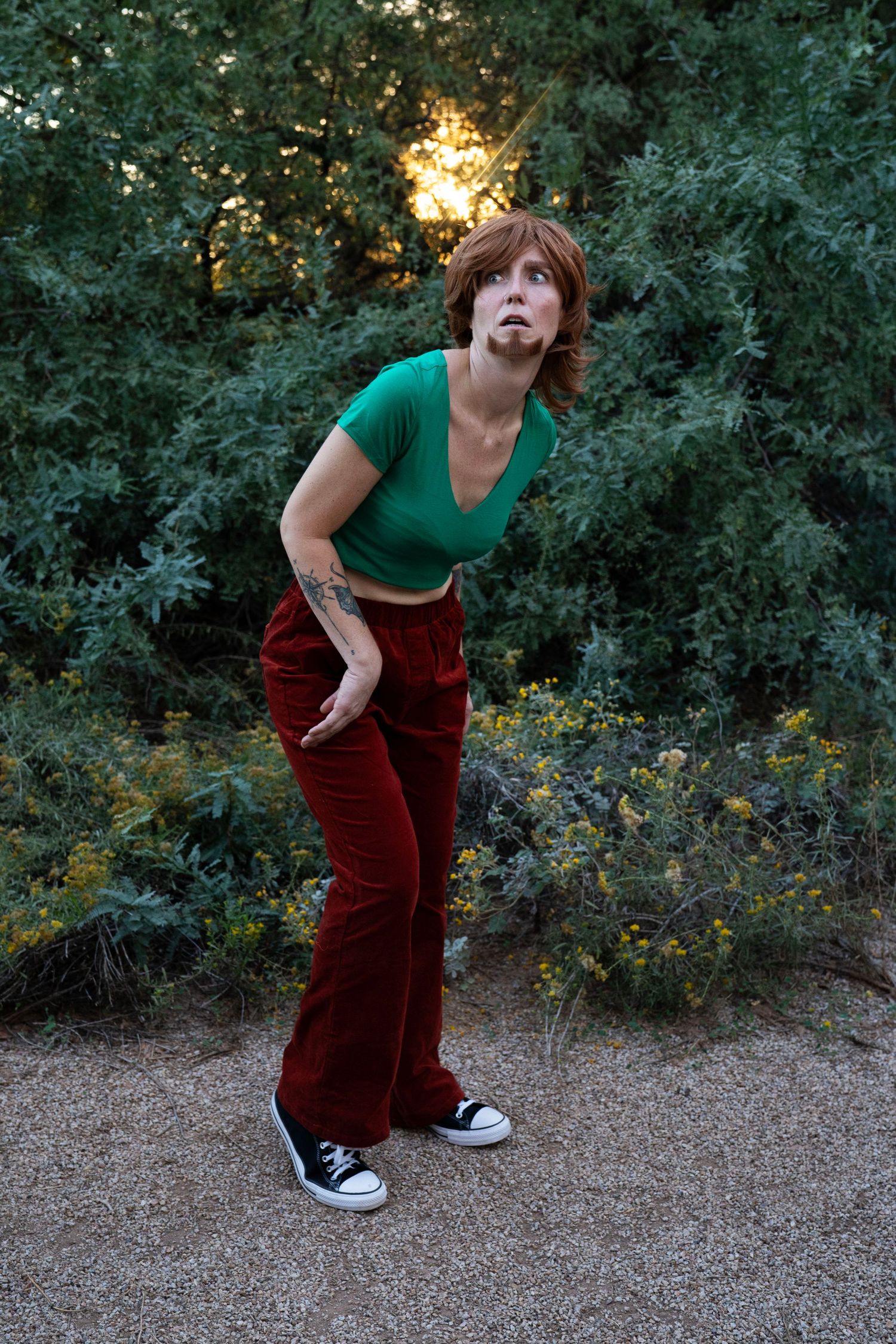 Person in a green crop top, red corduroy pants and black sneakers leaning forward in a playful pose on a gravel path by sunlit green shrubs.