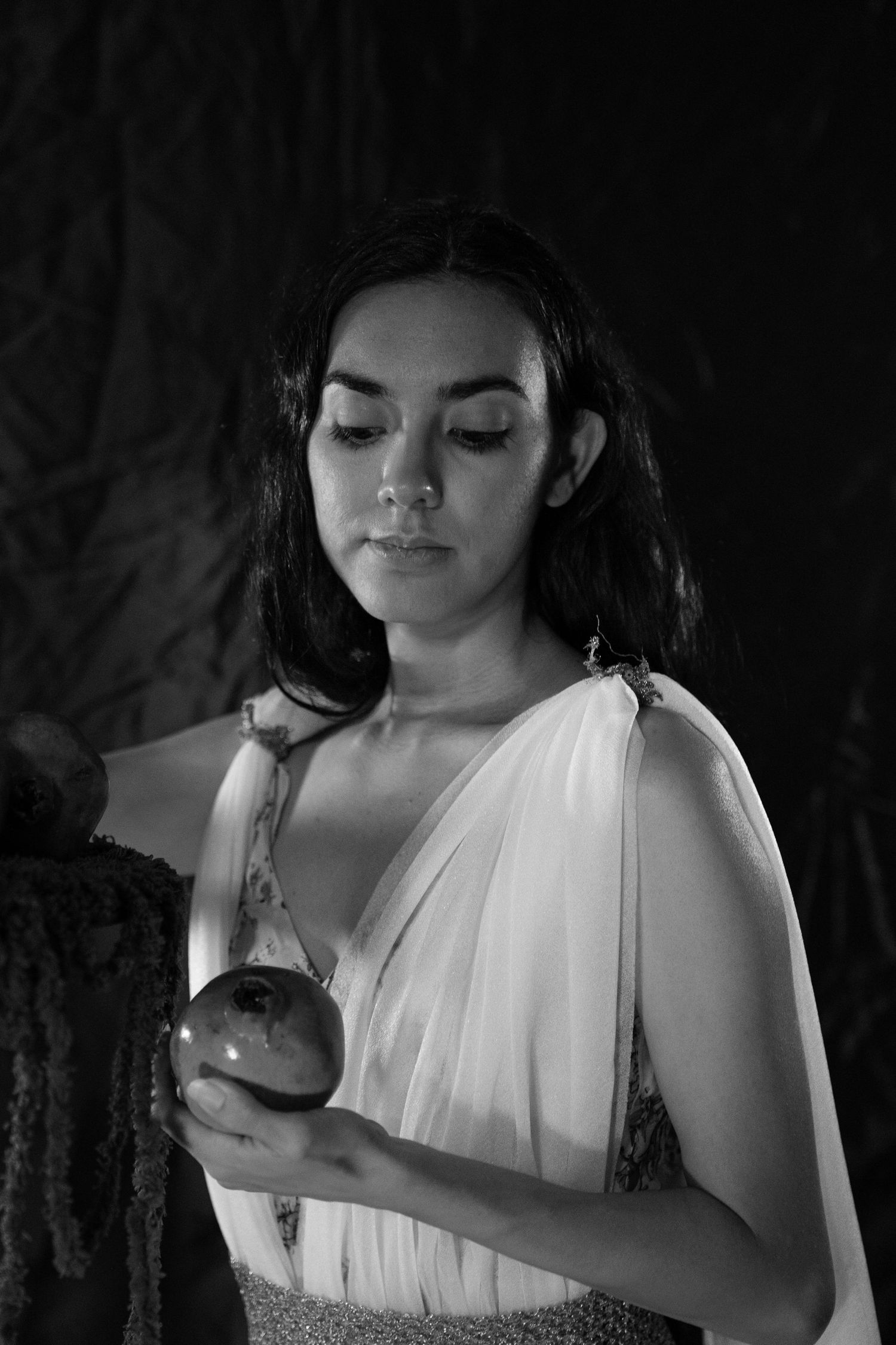 Black and white portrait of a woman in a flowing white draped gown holding a dark pomegranate, gazing downward in a dramatic setting.