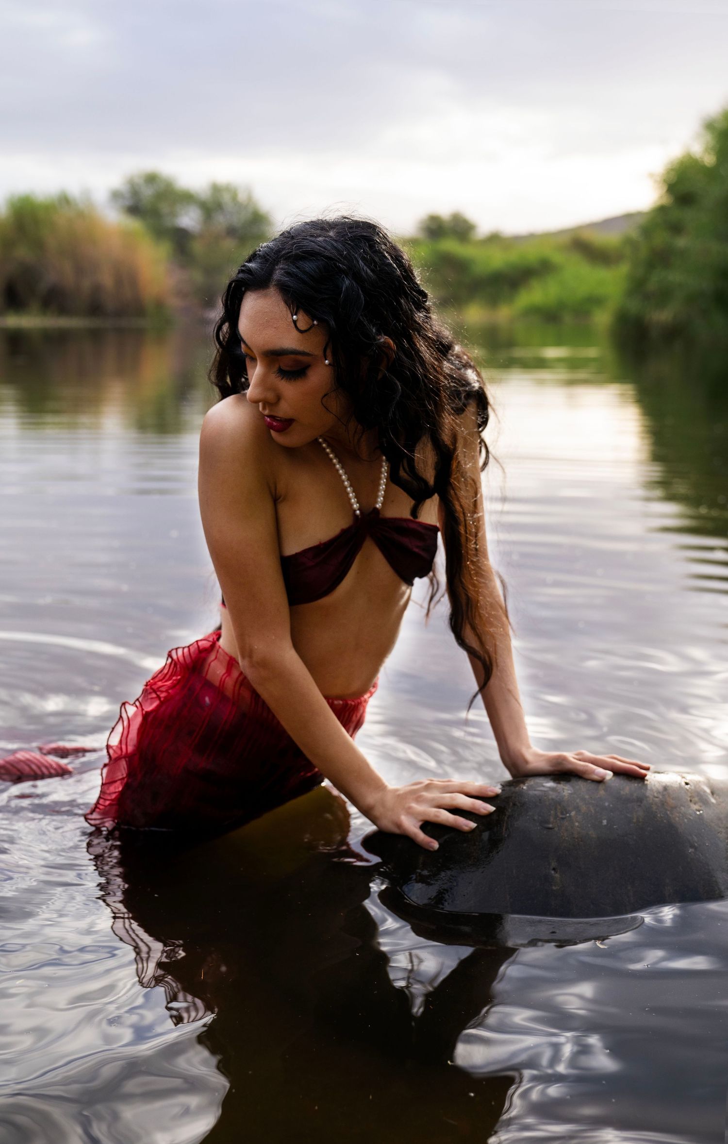 A dark-haired woman in a burgundy bralette and red skirt leans over a rock in a river under a cloudy sky.