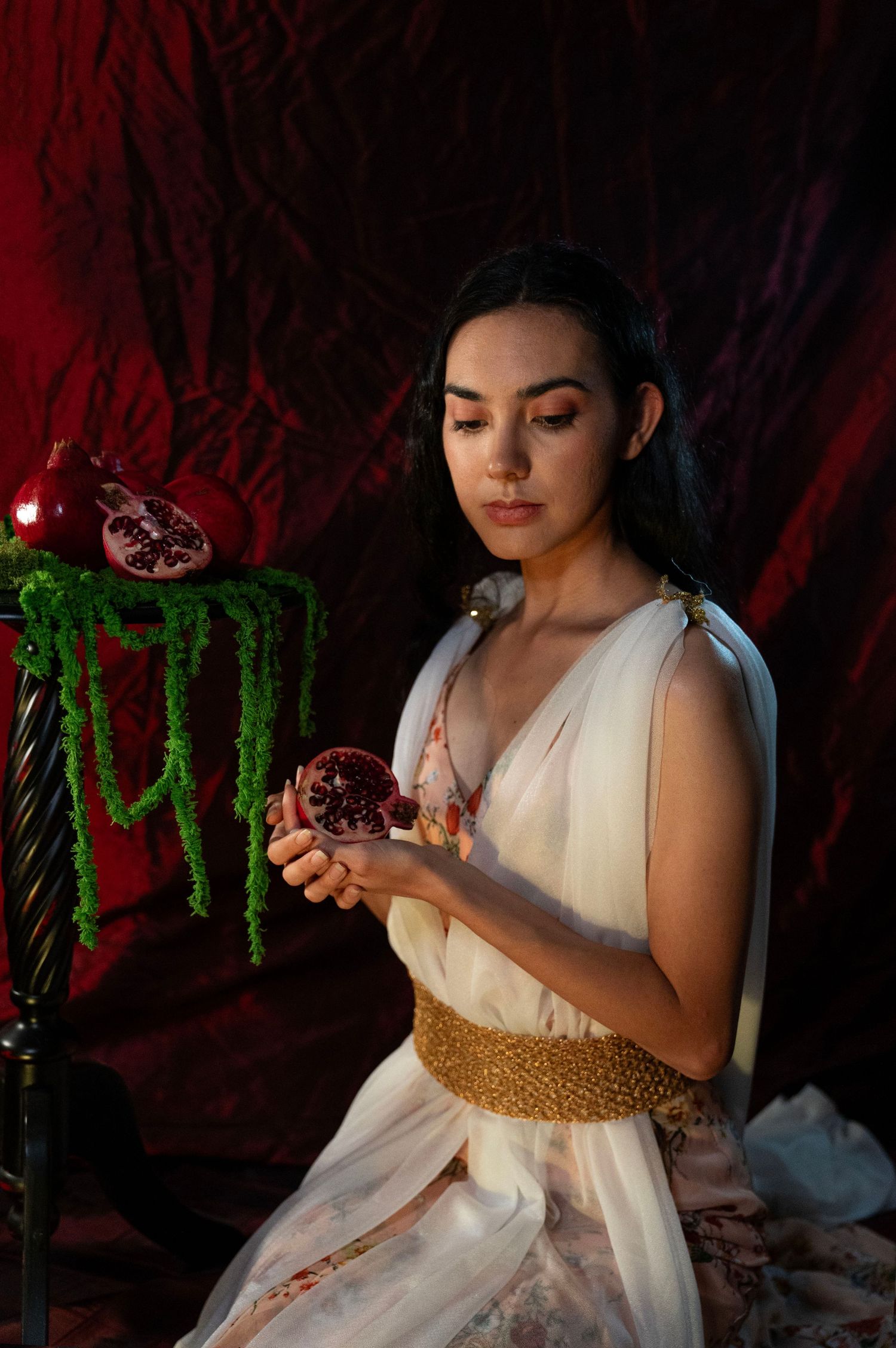 Woman in a flowing white gown and gold belt gazes down at a halved pomegranate beside green hanging amaranth on a dark red set.