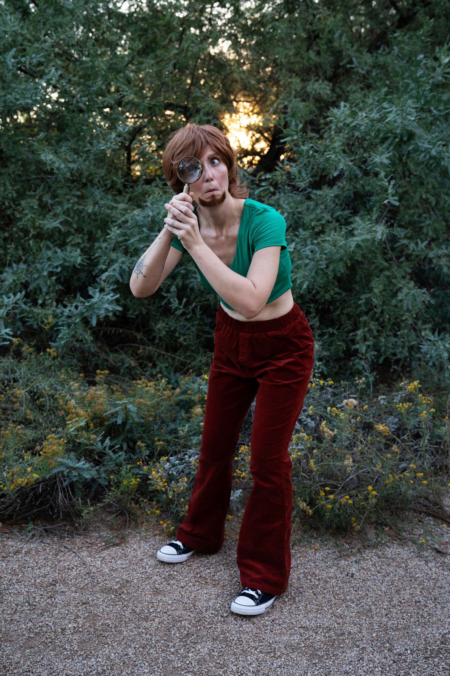 Woman in a green crop top, red corduroy flare pants and black sneakers peers through a magnifying glass in front of leafy bushes at sunset.
