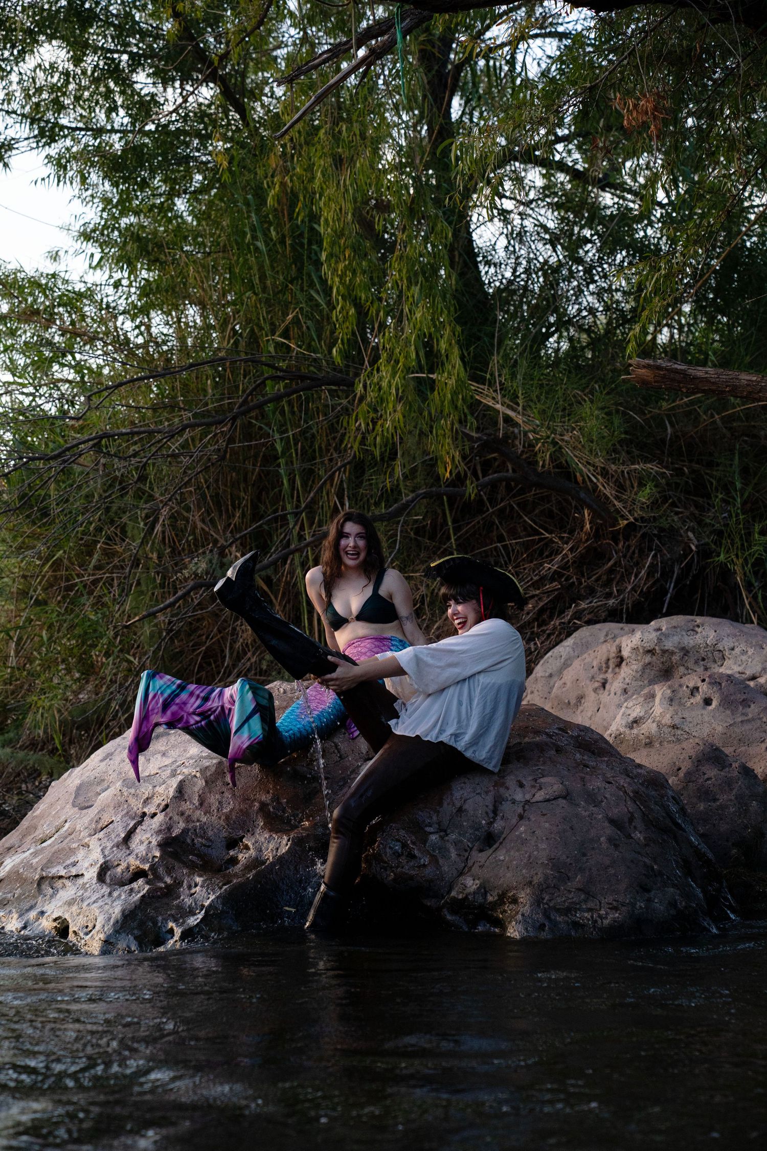 A smiling woman in a mermaid tail and a pirate-costumed person interact playfully on river rocks. Mermaid photoshoot, fantasy photoshoot.
