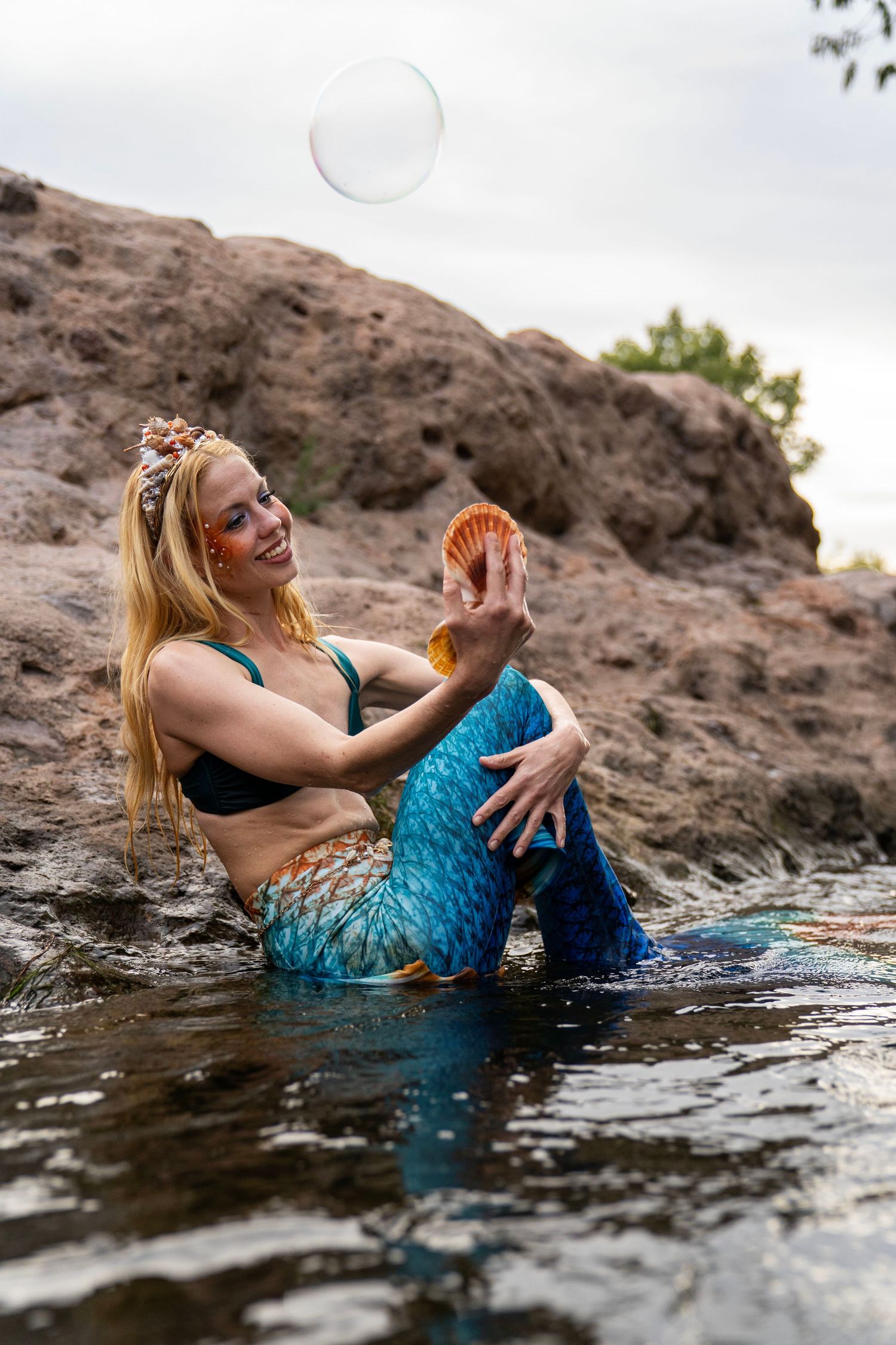A blonde woman in blue mermaid tail sits in shallow water holding a seashell with a bubble above. Fantasy Photoshoot