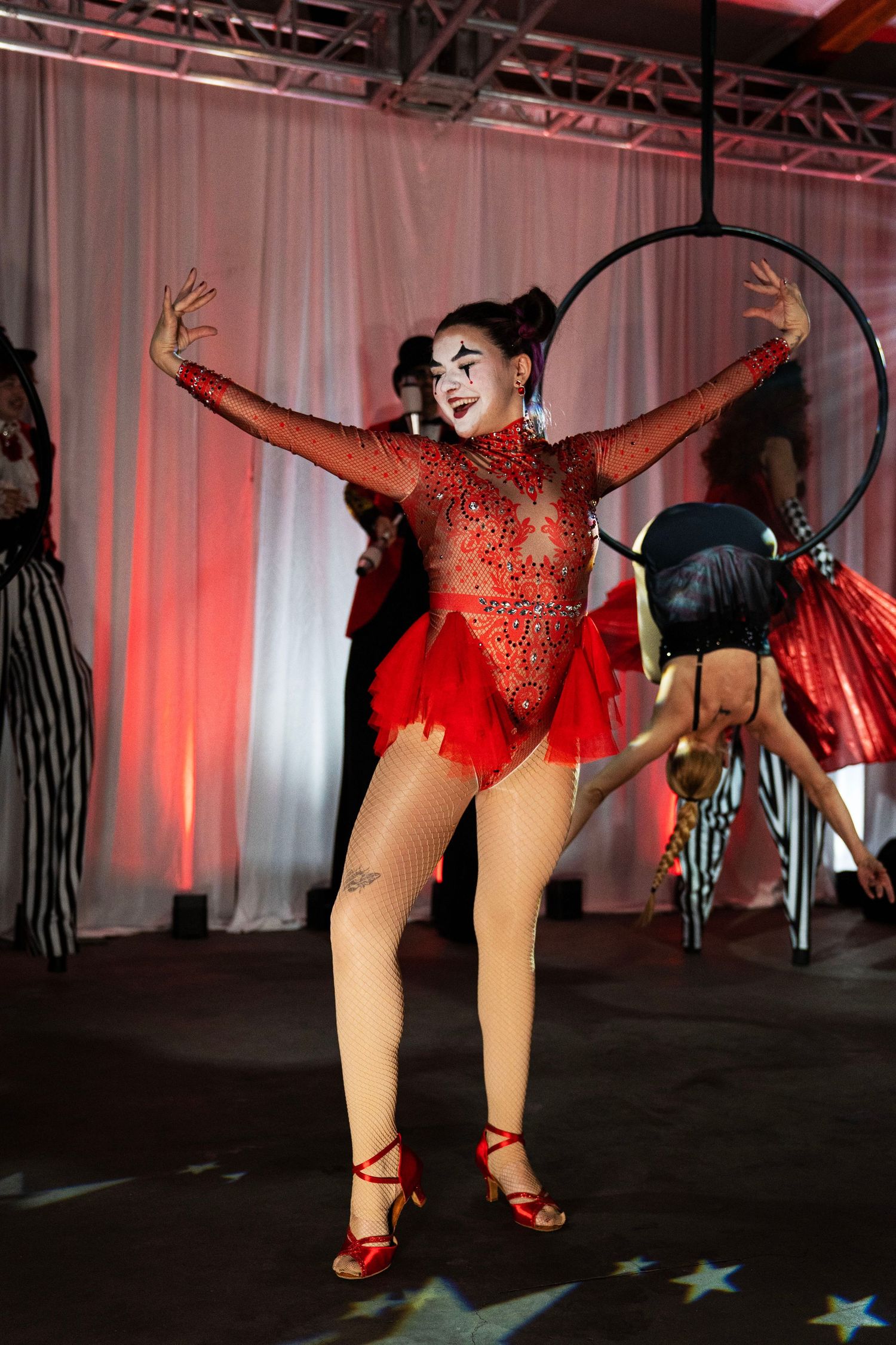 A circus performer in a red rhinestone bodysuit with clown makeup dances energetically on a star-lit stage.