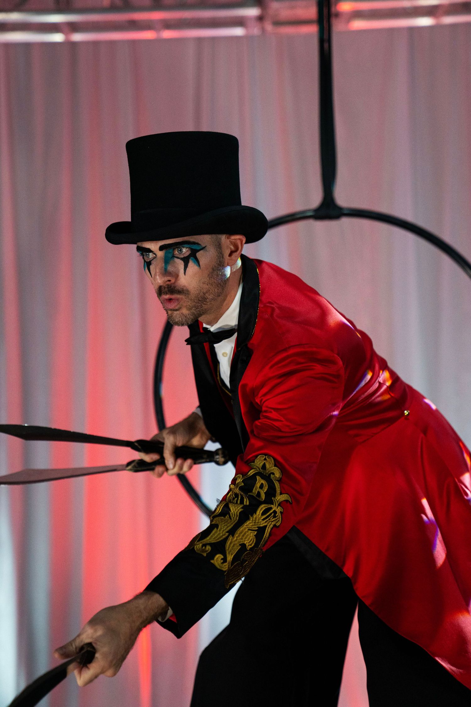 Male circus performer in red tailcoat and top hat with dramatic face paint holding aerial hoop props. Circus Photoshoot