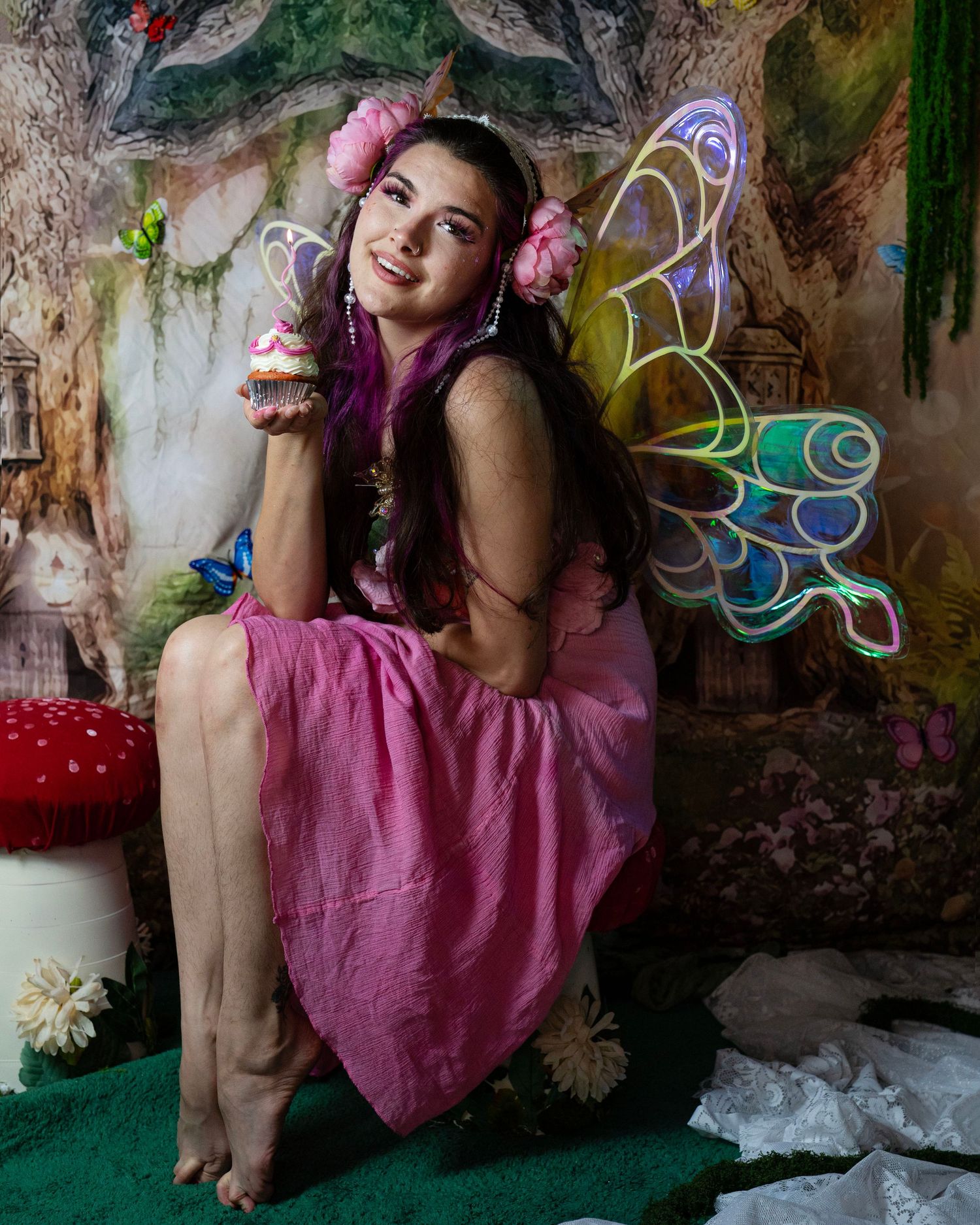 Young woman in pink fairy costume with colorful glowing wings and floral headpiece sits in enchanted forest studio. Fantasy Photoshoot