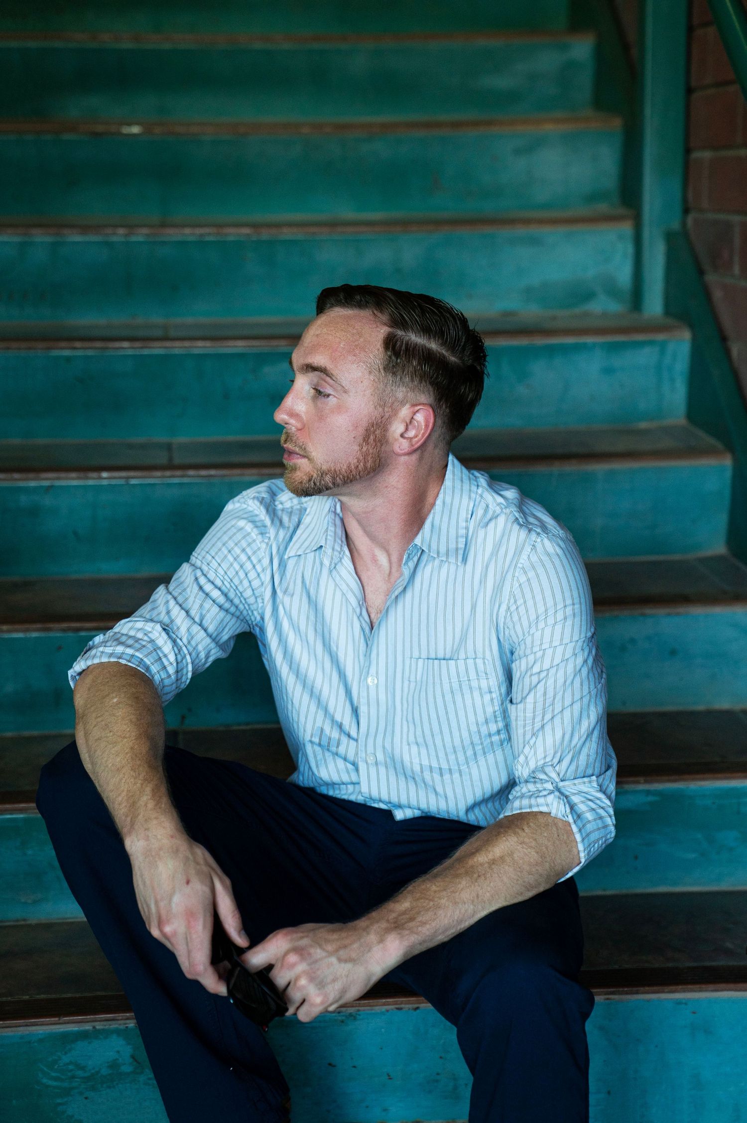 Young man in striped shirt sitting thoughtfully on turquoise vintage stairs outdoors.