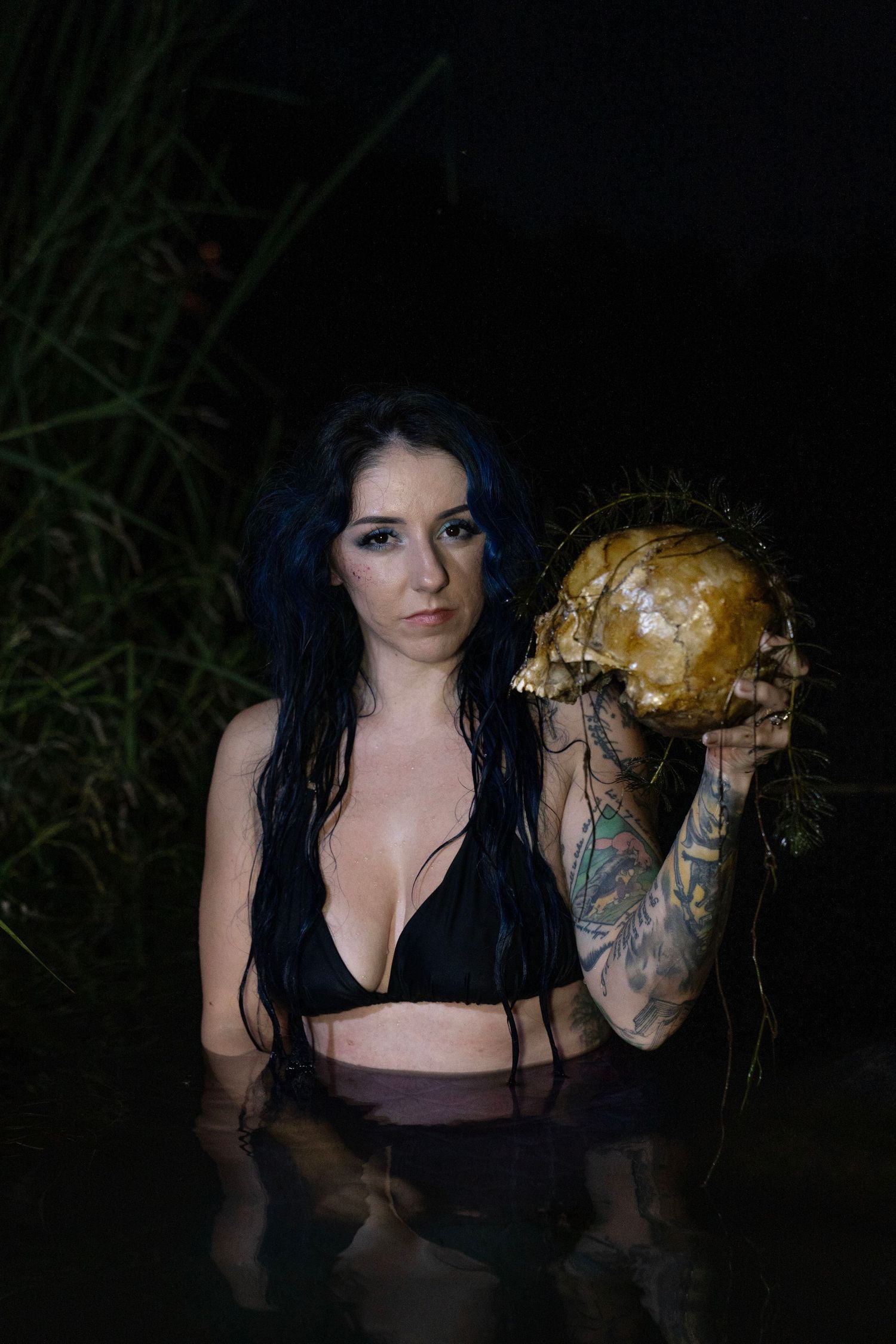 Blue-haired tattooed woman in black bikini holds skull while standing in dark water.