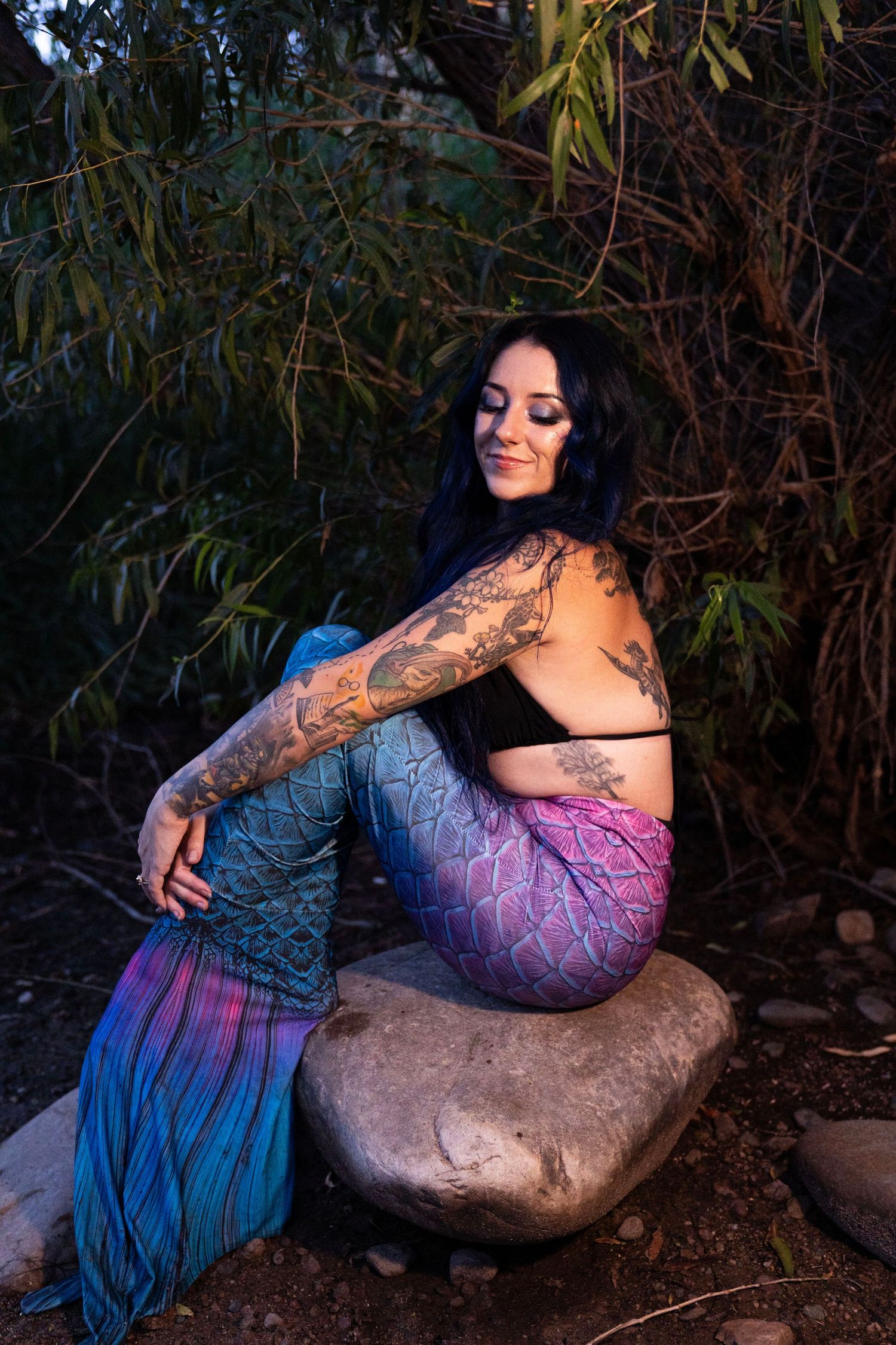 Tattooed woman in colorful mermaid tail costume sitting on a rock surrounded by dark foliage.
