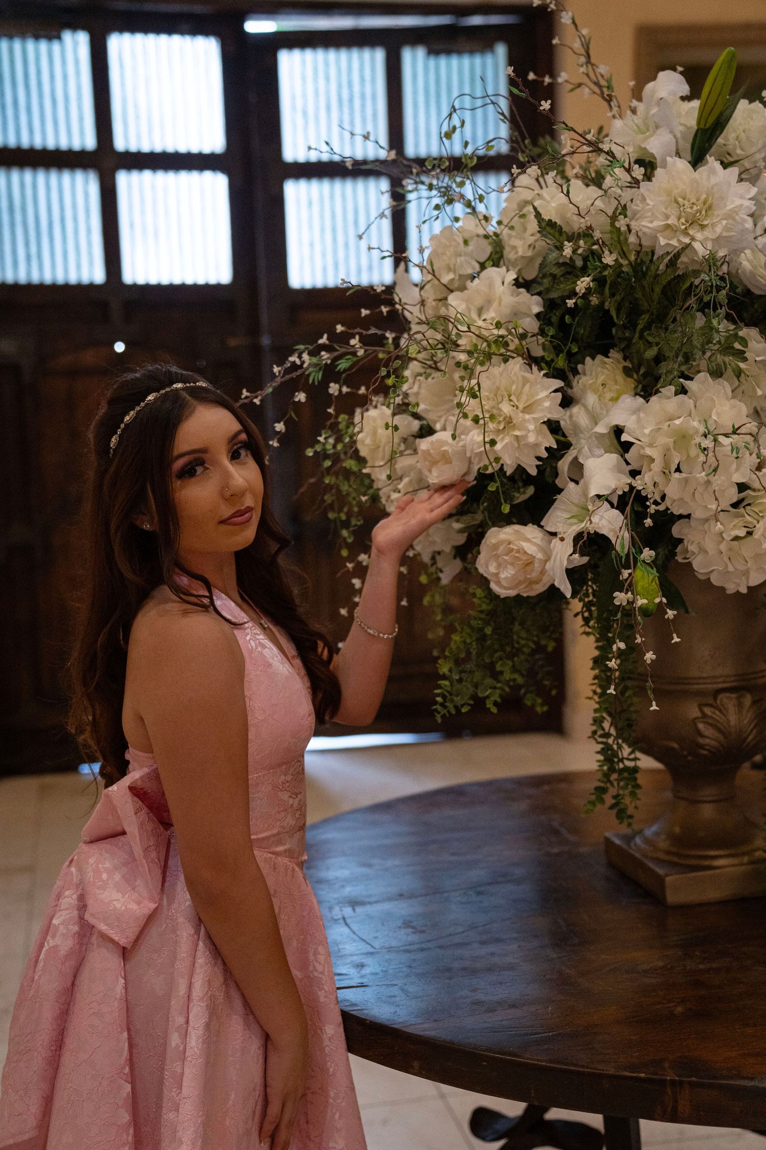 Young woman in pink gown touching large white floral arrangement on wooden table.