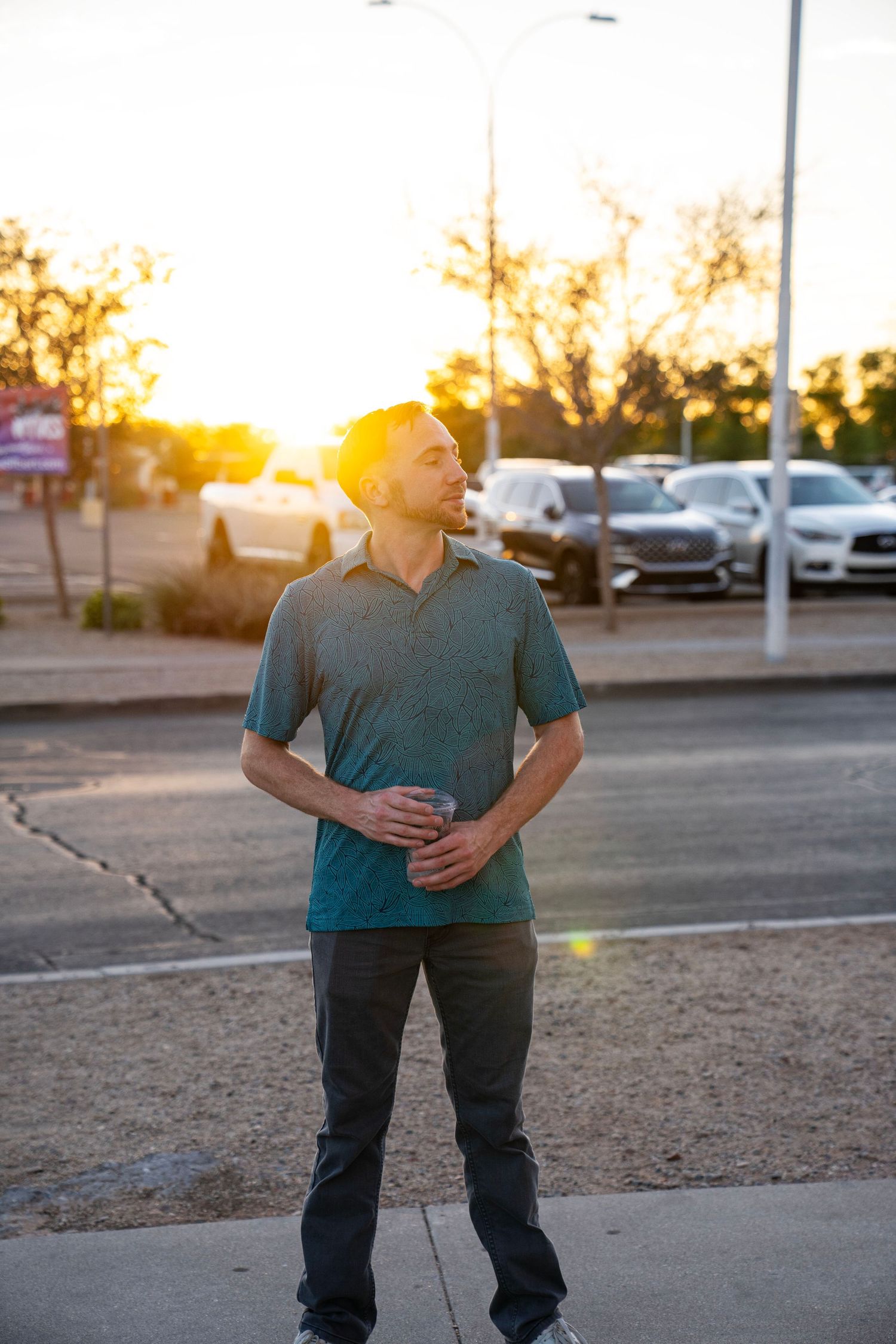 Man in teal polo holding a drink outdoors at golden hour sunset in a parking lot.