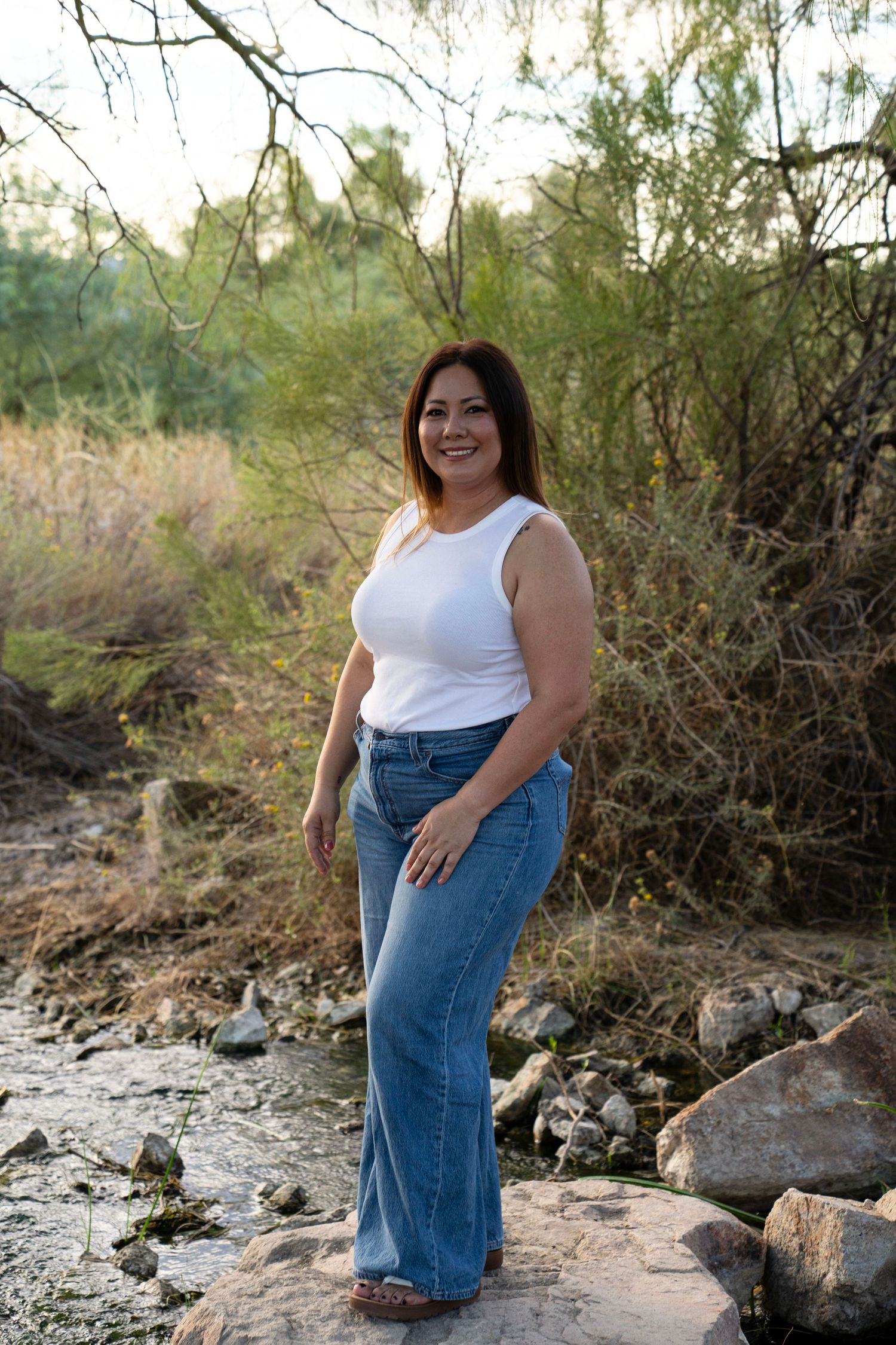 Woman in white tank top and wide-leg jeans smiling outdoors on rocks near a stream surrounded by trees.