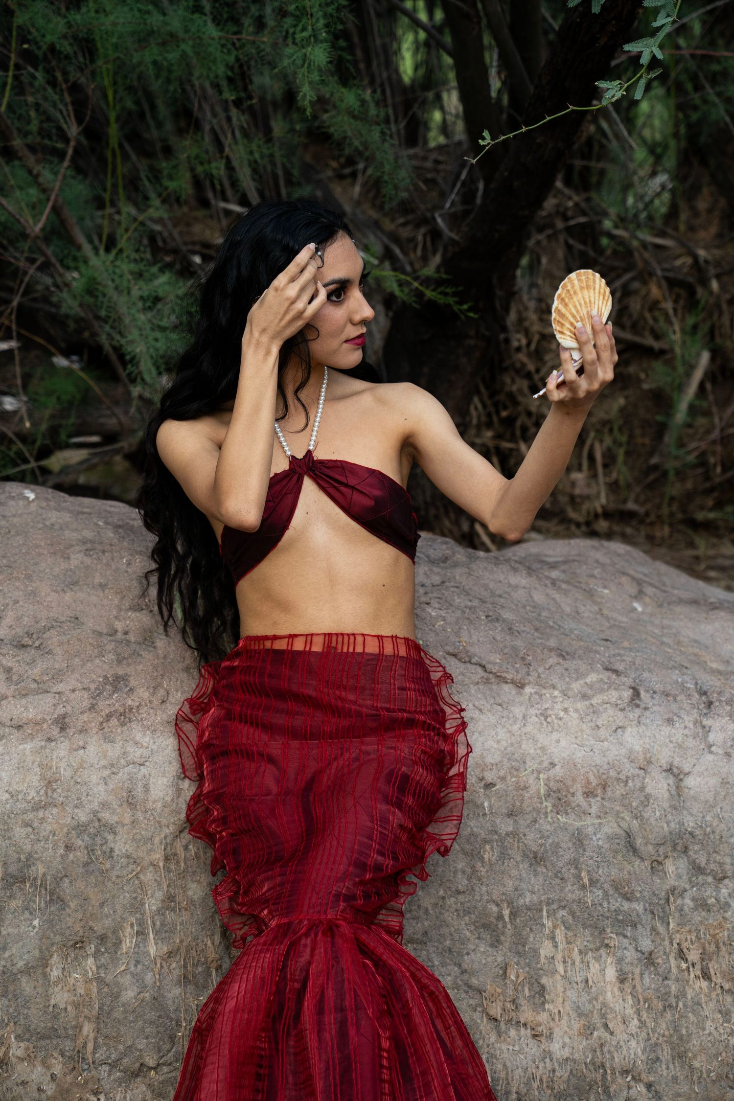Dark-haired woman in red mermaid tail and pearl necklace holding a scallop shell on rocks.