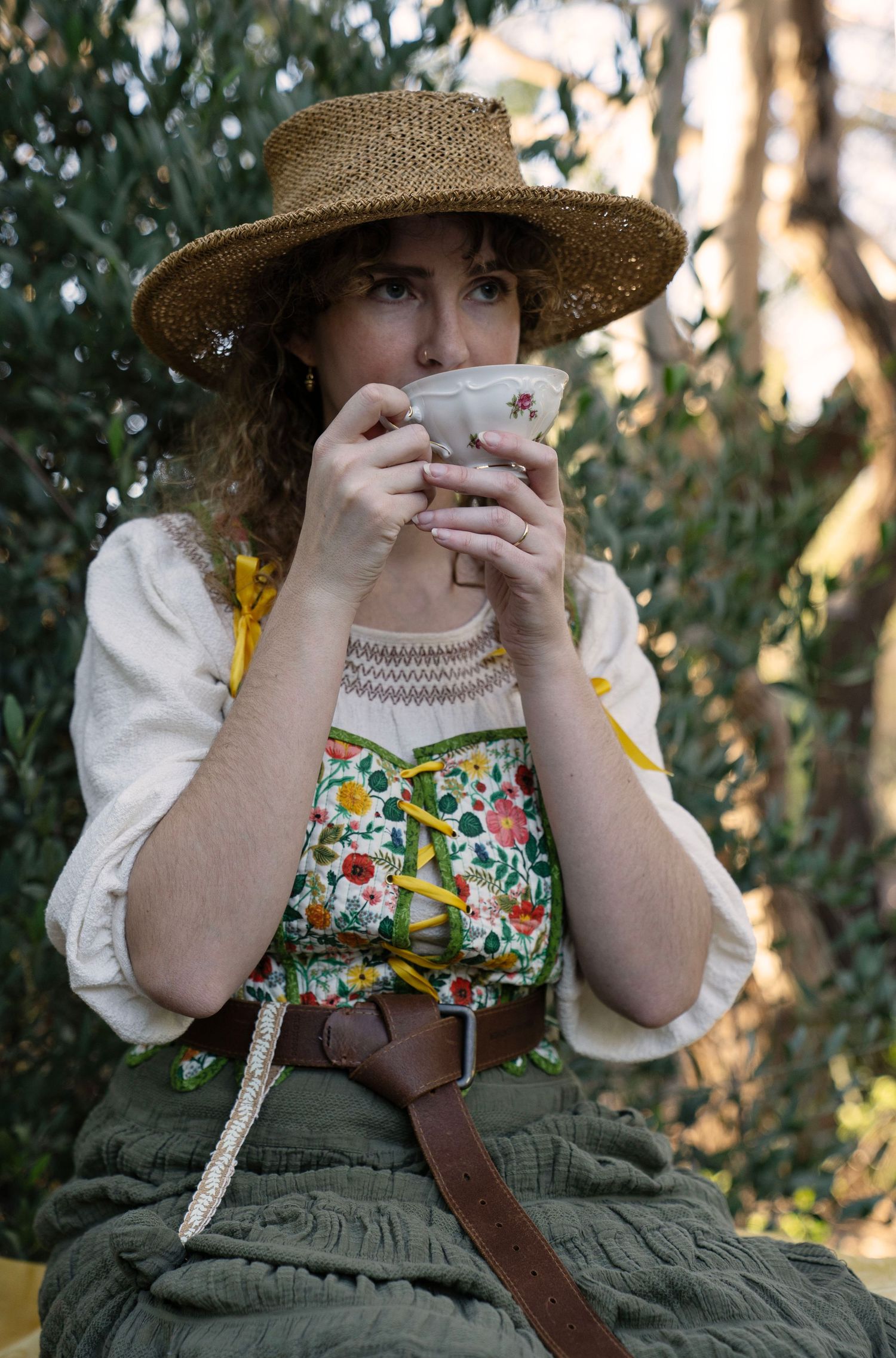 Woman in a straw hat and floral bodice with green skirt sips tea from a delicate china cup while seated outdoors among leafy trees.