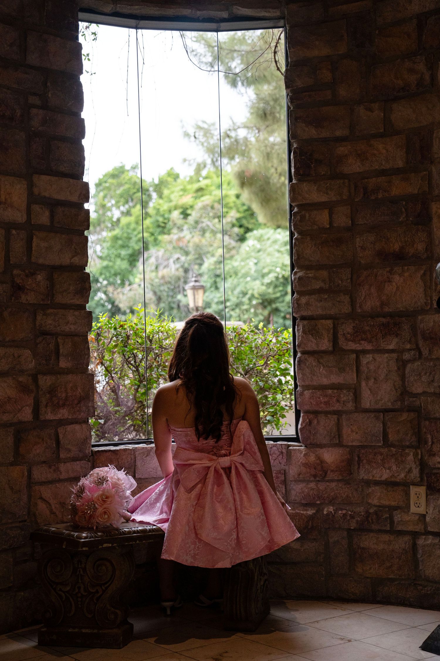 Girl in pink bow dress gazing out stone castle window with garden view.