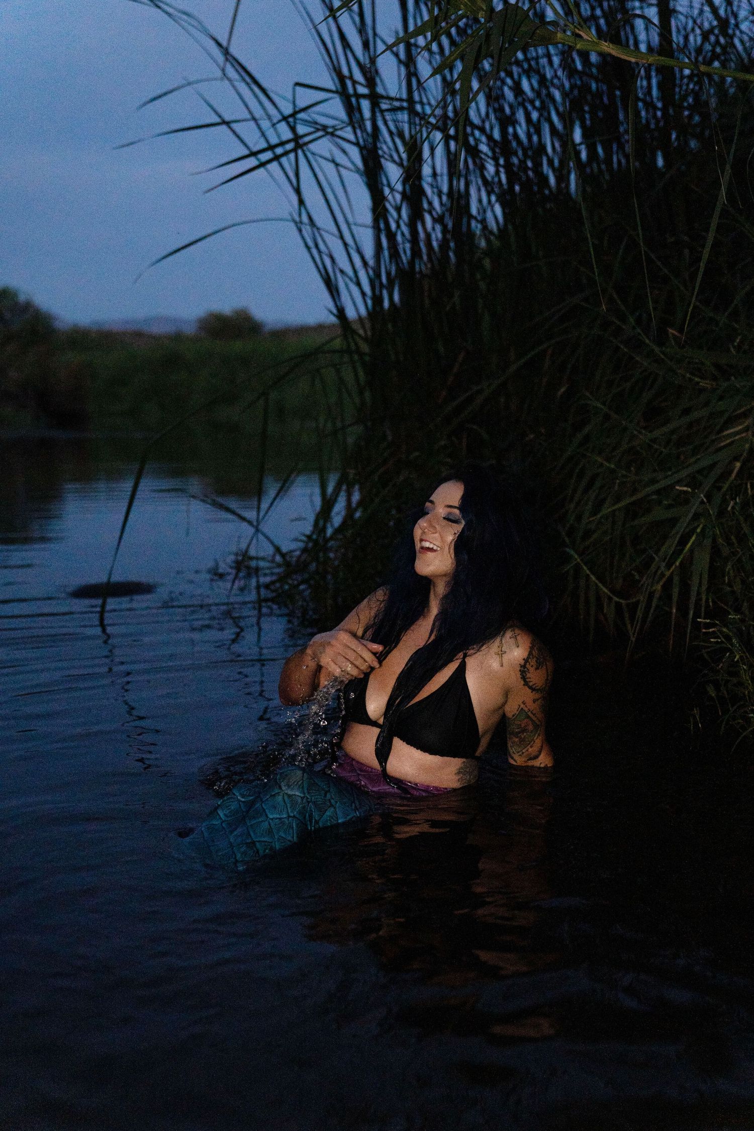 Tattooed woman in mermaid costume laughing while submerged in a dark river among tall reeds.