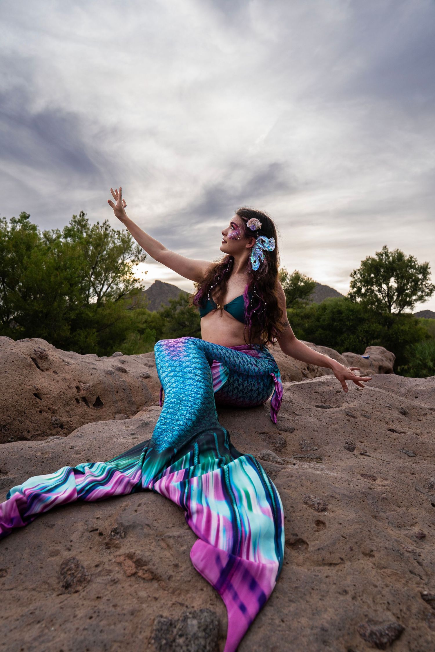 Woman in a vibrant teal and purple mermaid tail with shell hair accessories posing on rocks outdoors. Fantasy Photoshoot