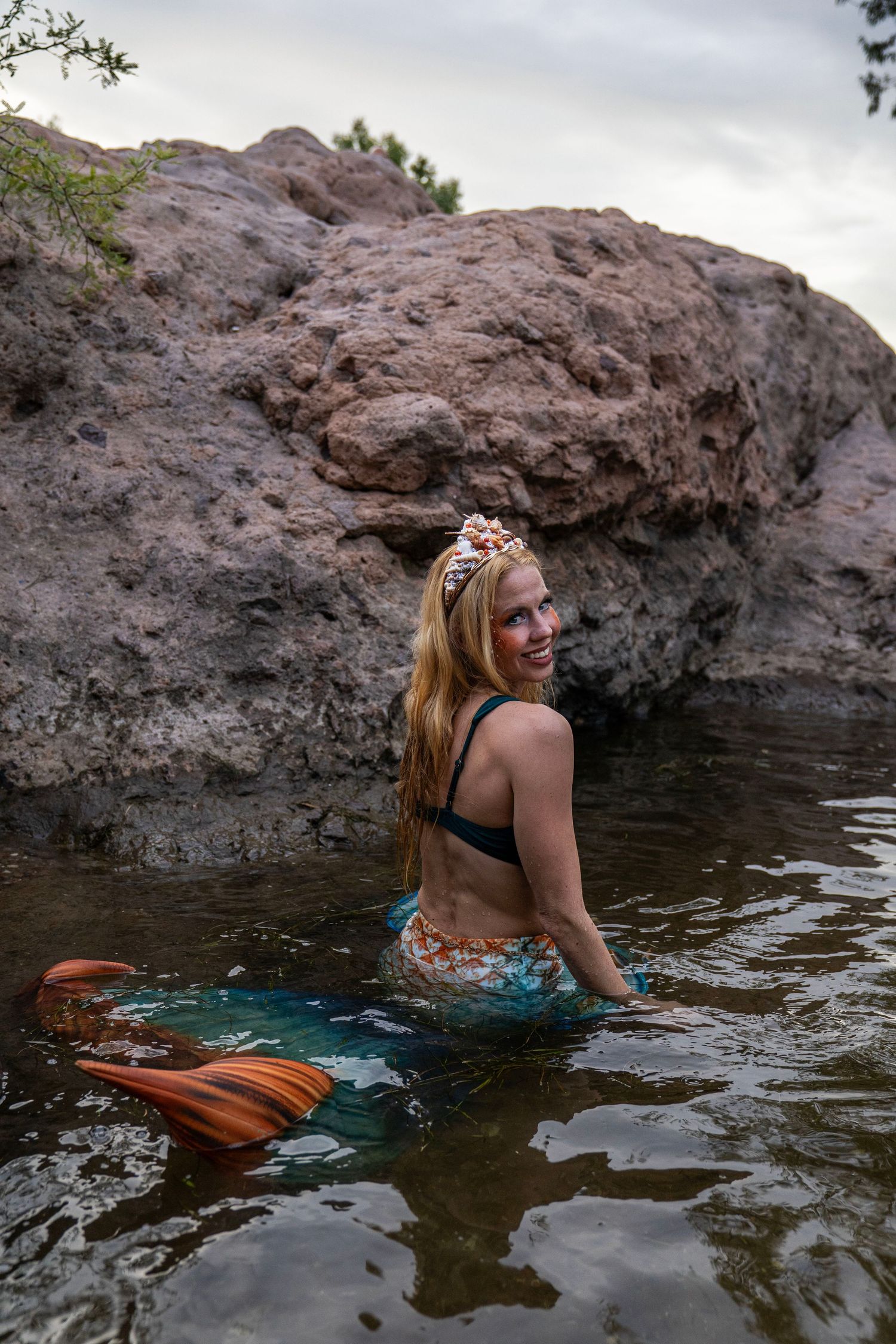Smiling woman in mermaid costume with orange tail and floral crown posing in rocky natural water. Fantasy Photoshoot