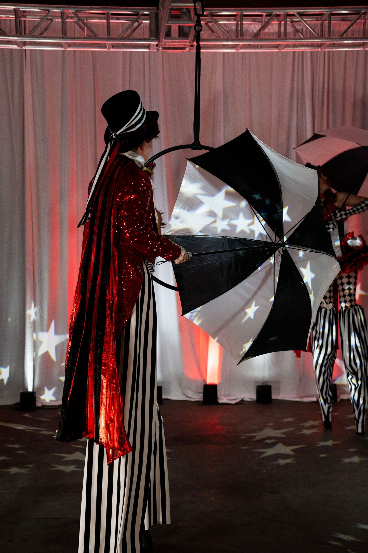Circus stilt walker in red sequin coat and striped pants holding black and white umbrella on stage.