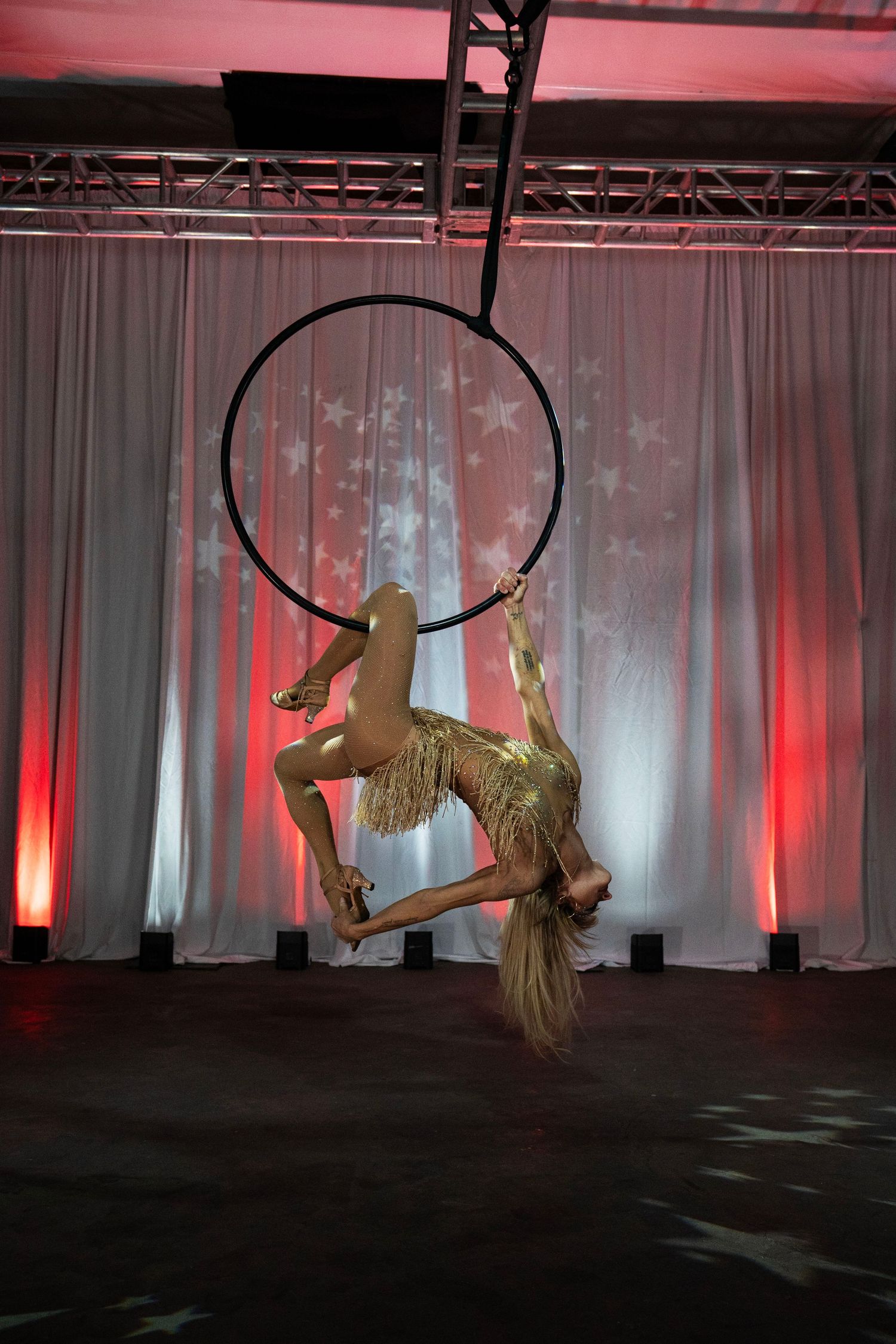 Female aerialist in gold costume performs a graceful split pose on a suspended lyra hoop against a star backdrop.