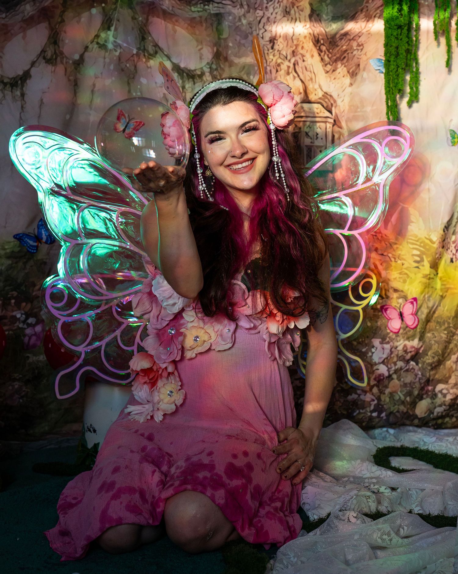 Smiling woman in pink floral fairy dress with glowing wings poses in a magical enchanted forest set. Fantasy Photoshoot