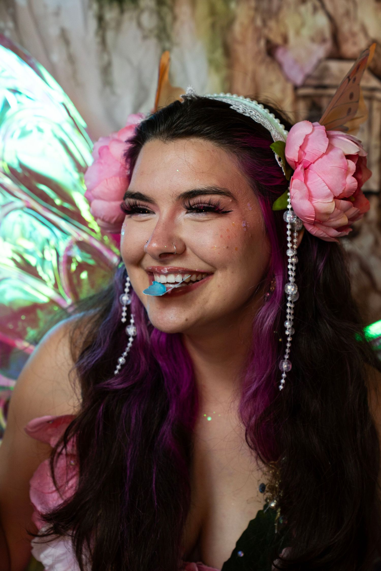 Smiling woman in fairy costume with pink flowers, purple hair, pearl earrings, and blue butterfly prop in mouth. Fantasy Photoshoot