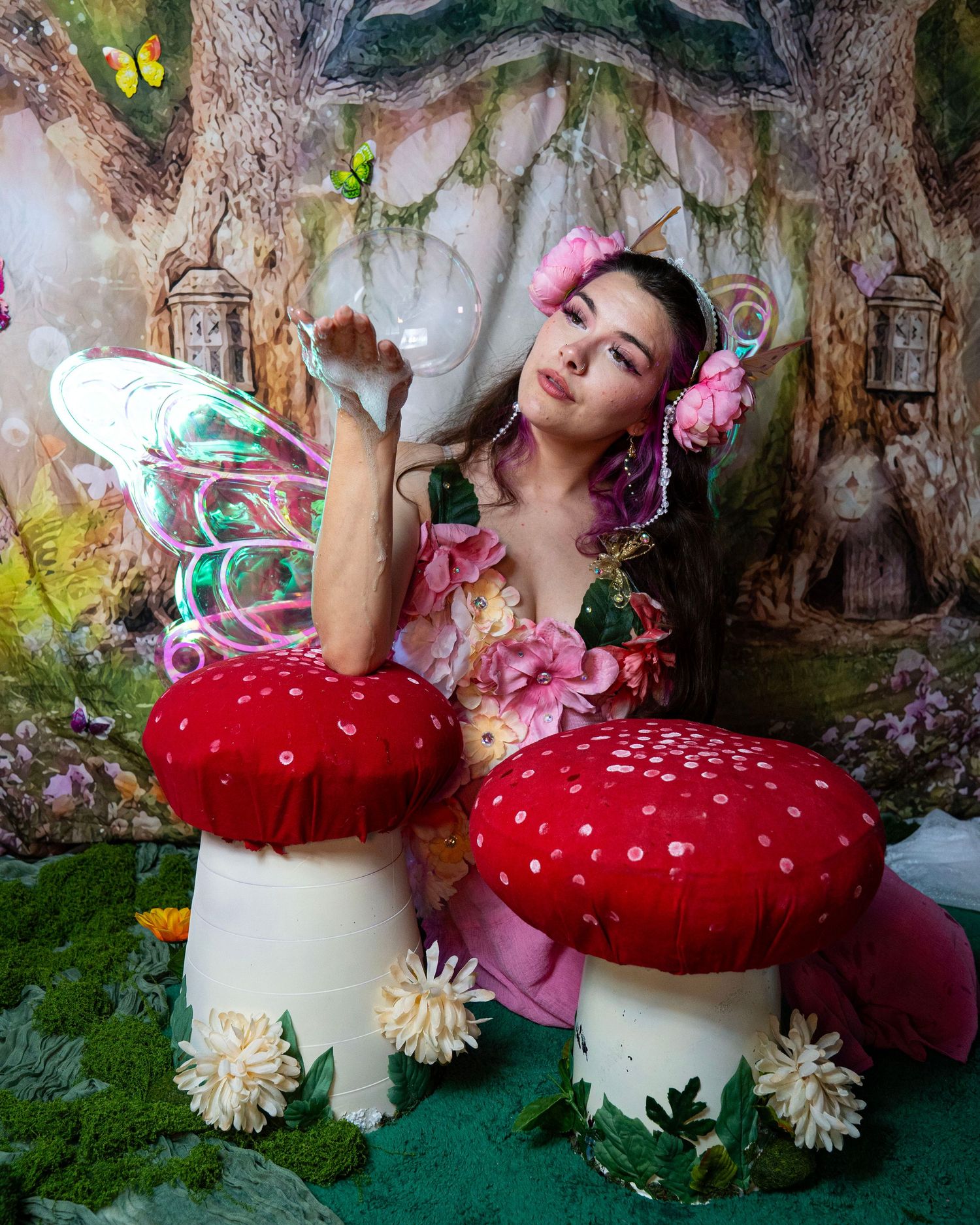 Fairy cosplay model with butterfly wings poses beside giant red spotted mushrooms in magical woodland studio set. Fantasy Photoshoot