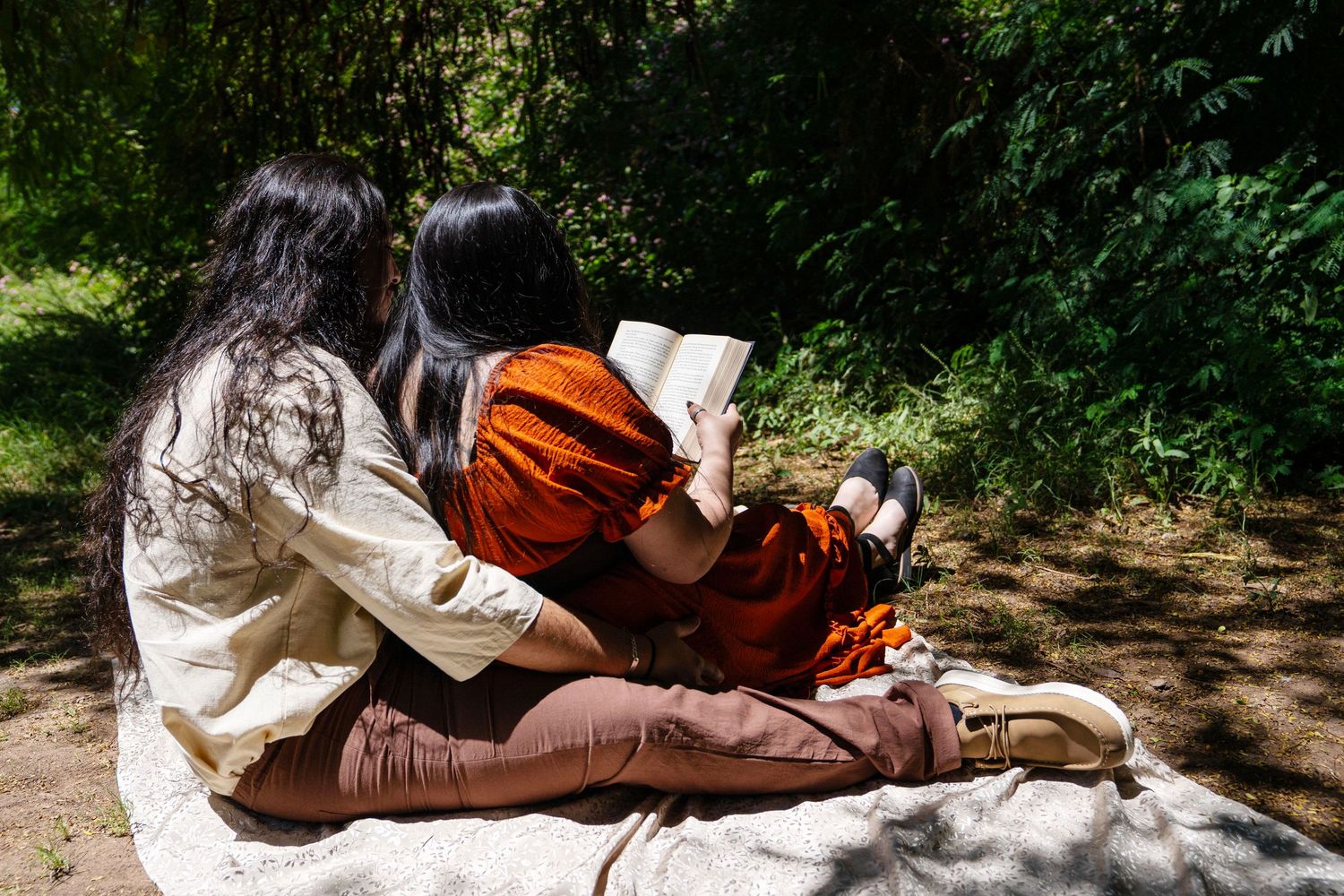 Two women with dark hair sitting on blanket outdoors reading a book surrounded by green foliage.