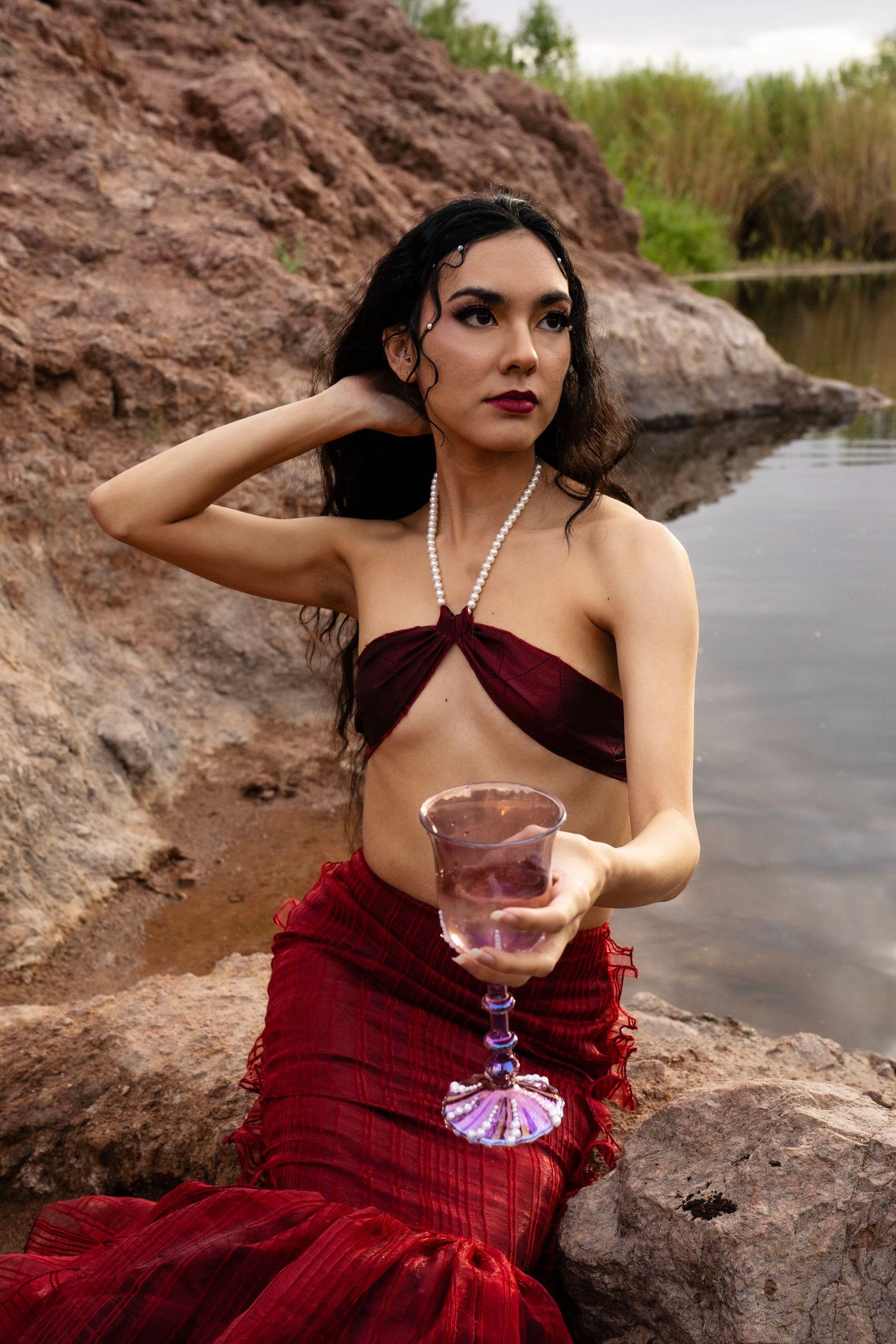 A woman in a red mermaid outfit sits on rocks by a river, holding a decorative pink glass with one hand raised.