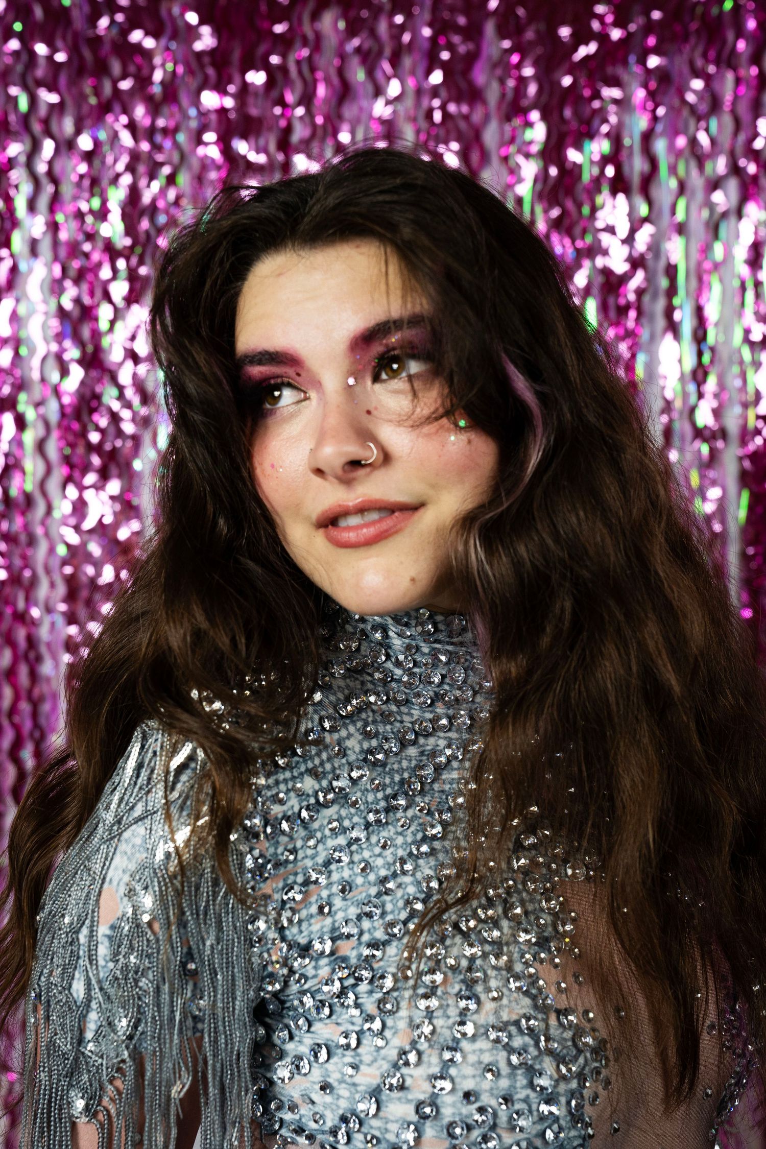 Young woman with long brunette hair and bold pink eye makeup wears silver rhinestone sheer top.