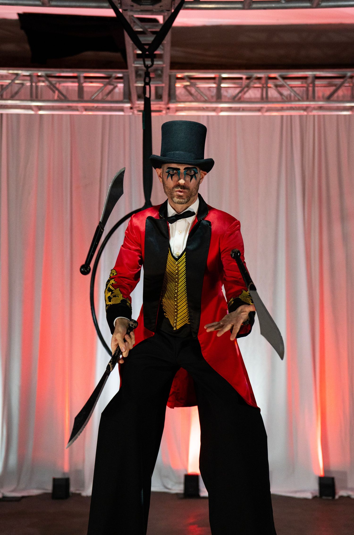 Male circus ringmaster in red tailcoat and top hat performs on stilts holding juggling knives.