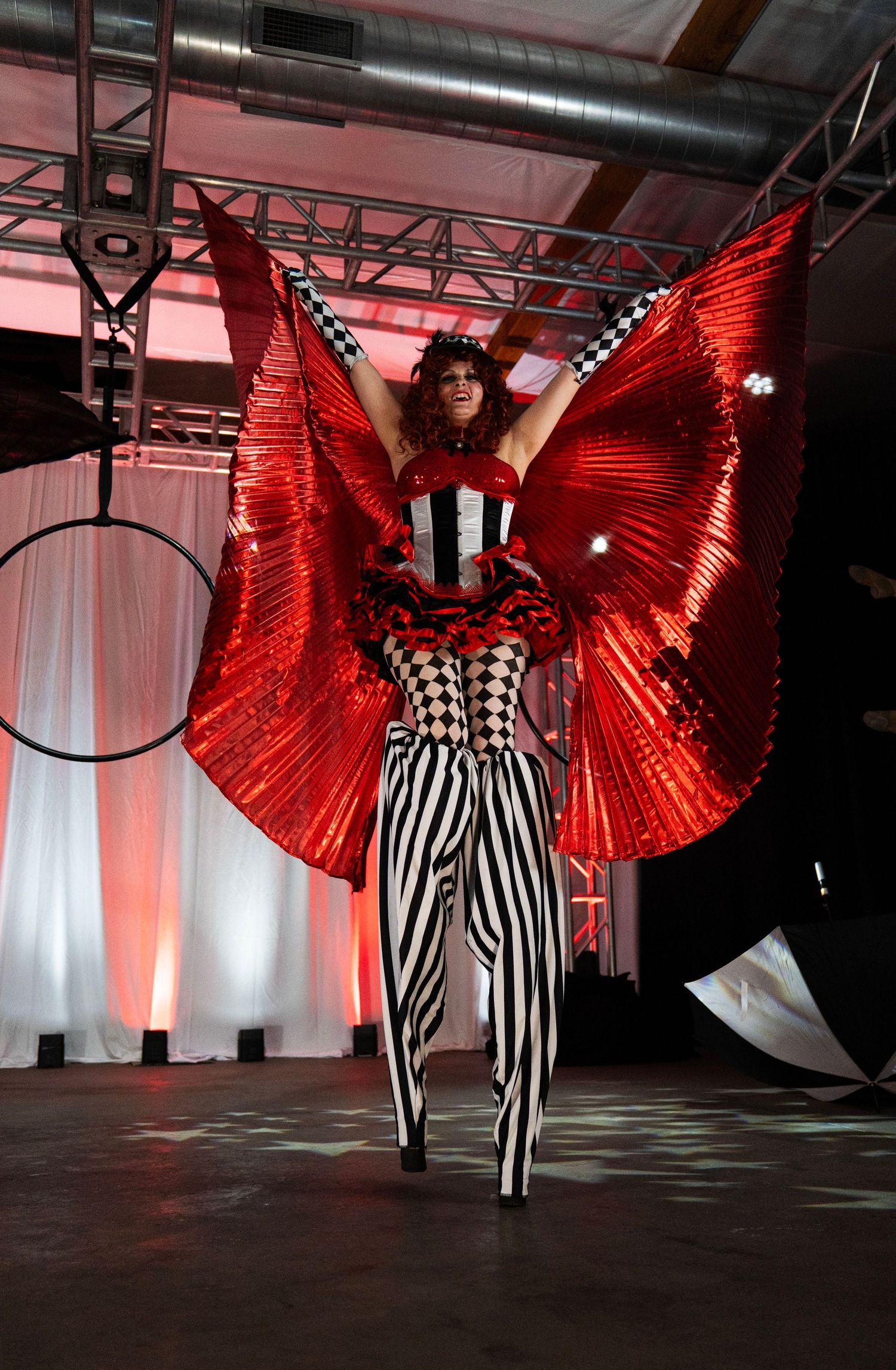 Female circus performer in harlequin corset raises red butterfly wings beside aerial hoop. Circus Photoshoot