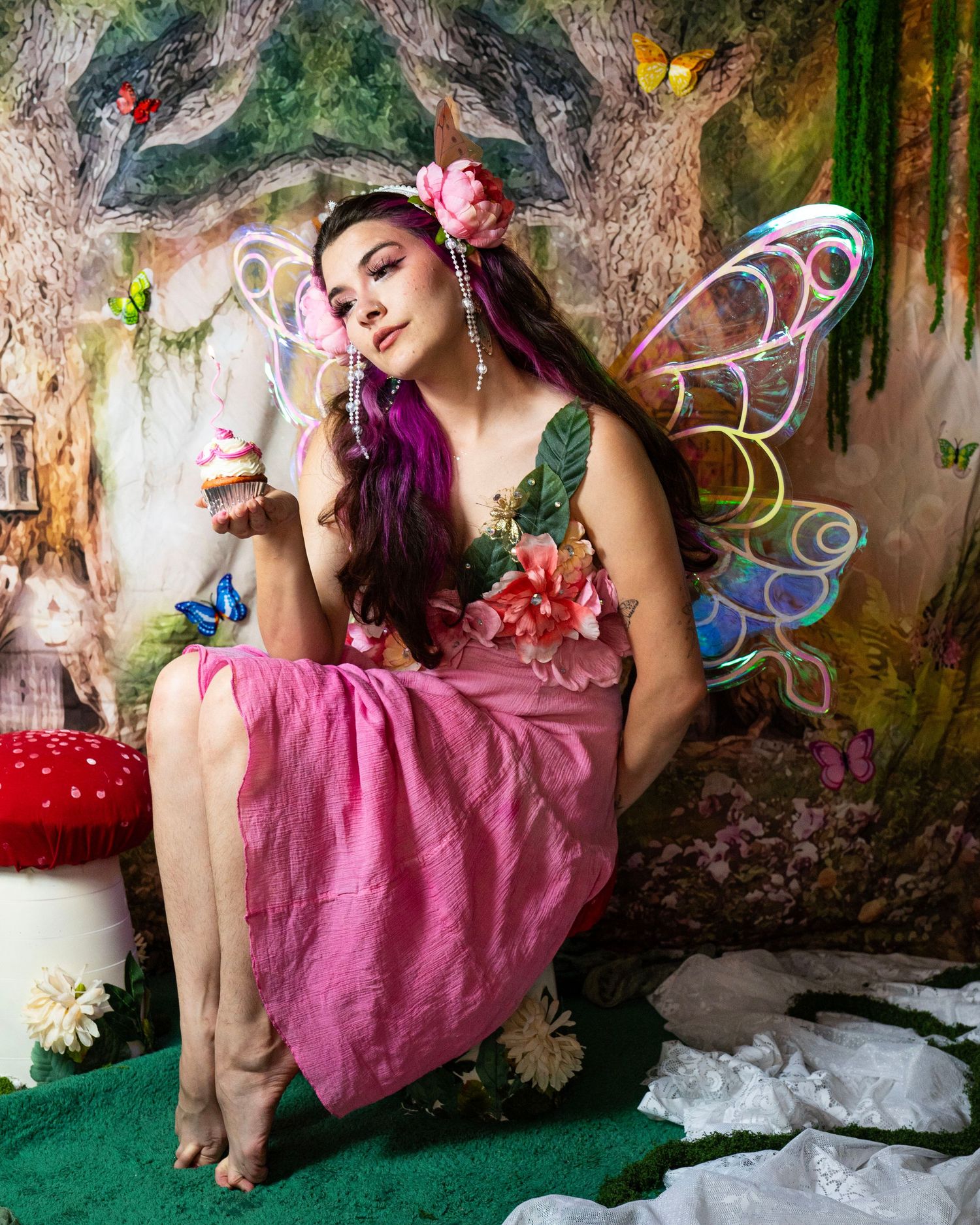 Woman in fairy costume with large floral bodice and rainbow wings holds cupcake in enchanted forest set. Fantasy Photoshoot