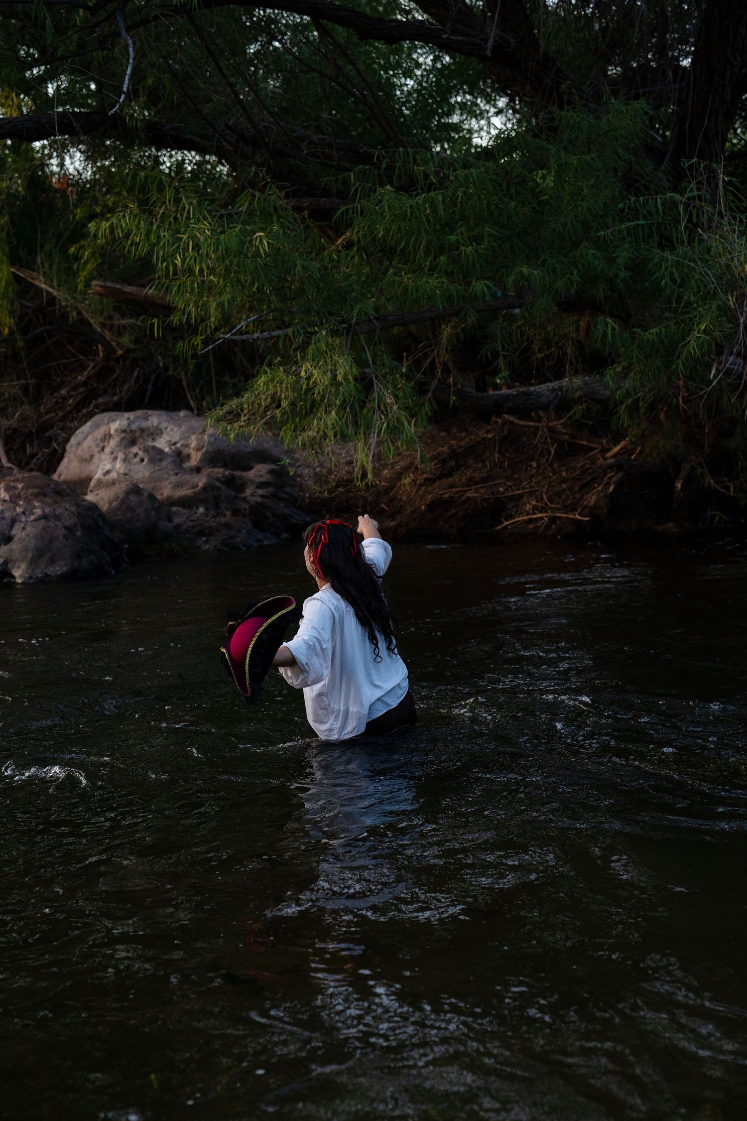 A person in a white shirt wades through a shallow river near rocky shores and dense green trees. Mermaid photoshoot, fantasy photoshoot.