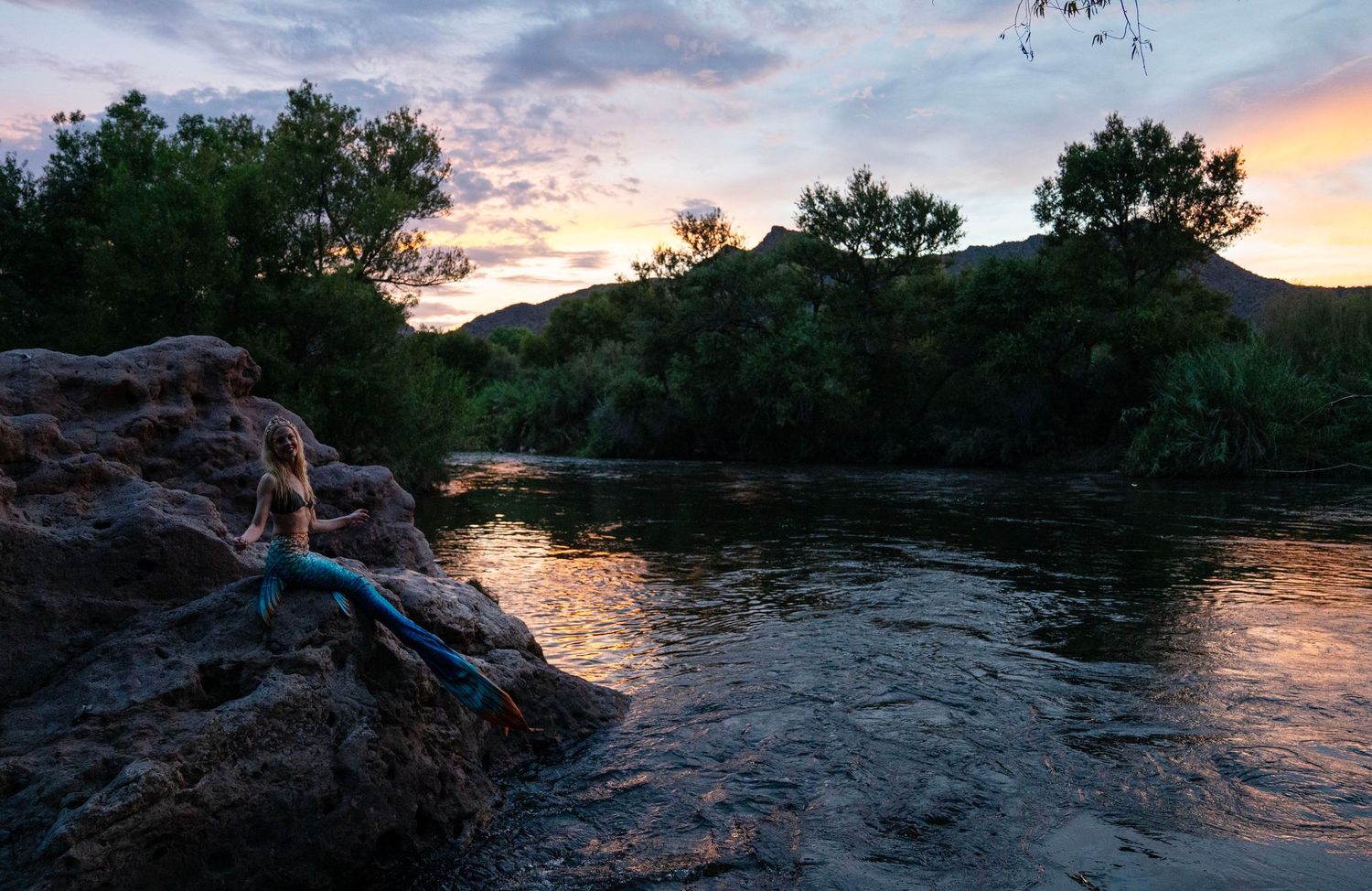 Woman in mermaid costume resting on rocks beside a river at a colorful sunset. Mermaid photoshoot, fantasy photoshoot. Arizona photographer.