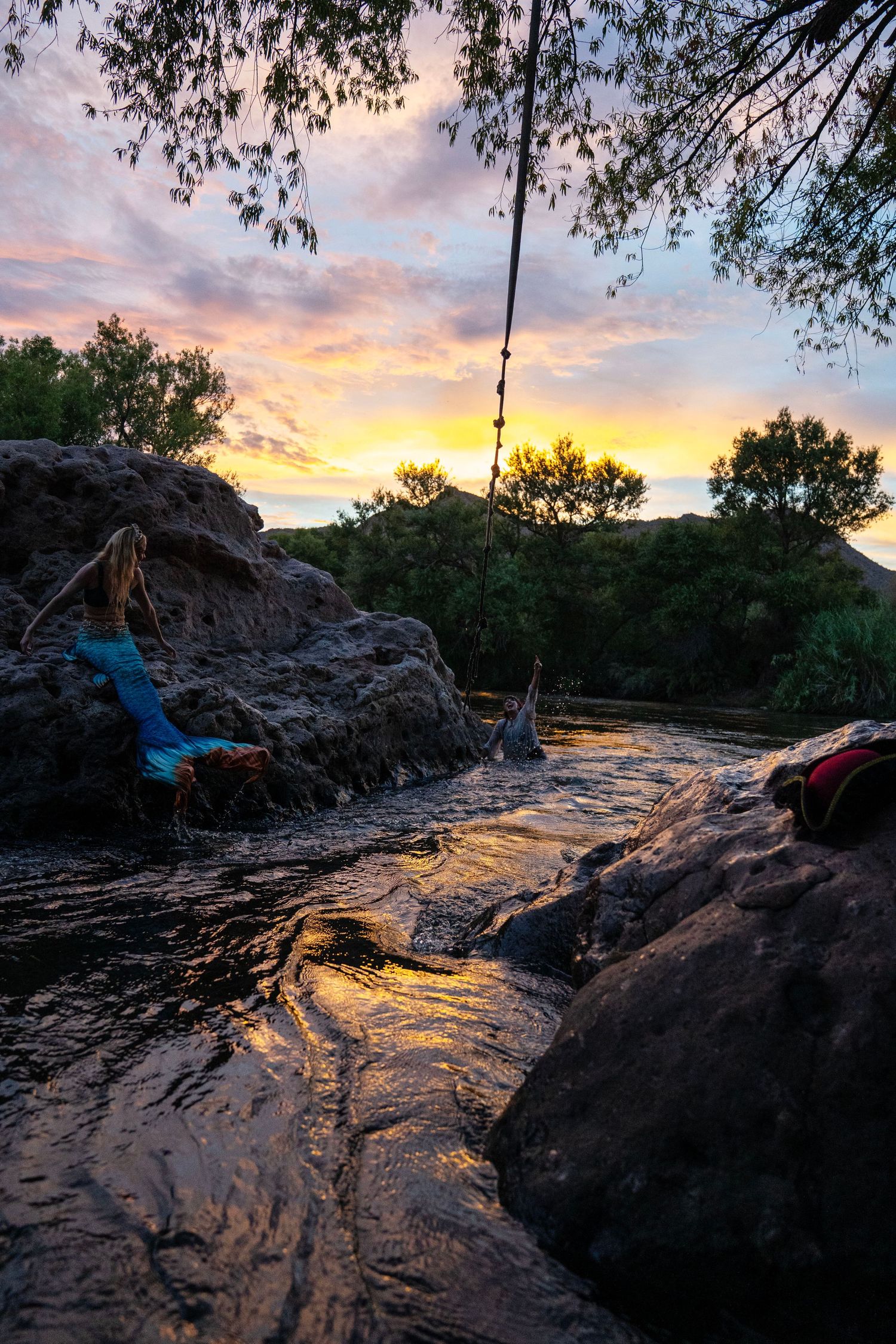 Person in mermaid tail on rocks near river with vibrant sunset sky. Mermaid photoshoot, fantasy photoshoot. Arizona photographer.