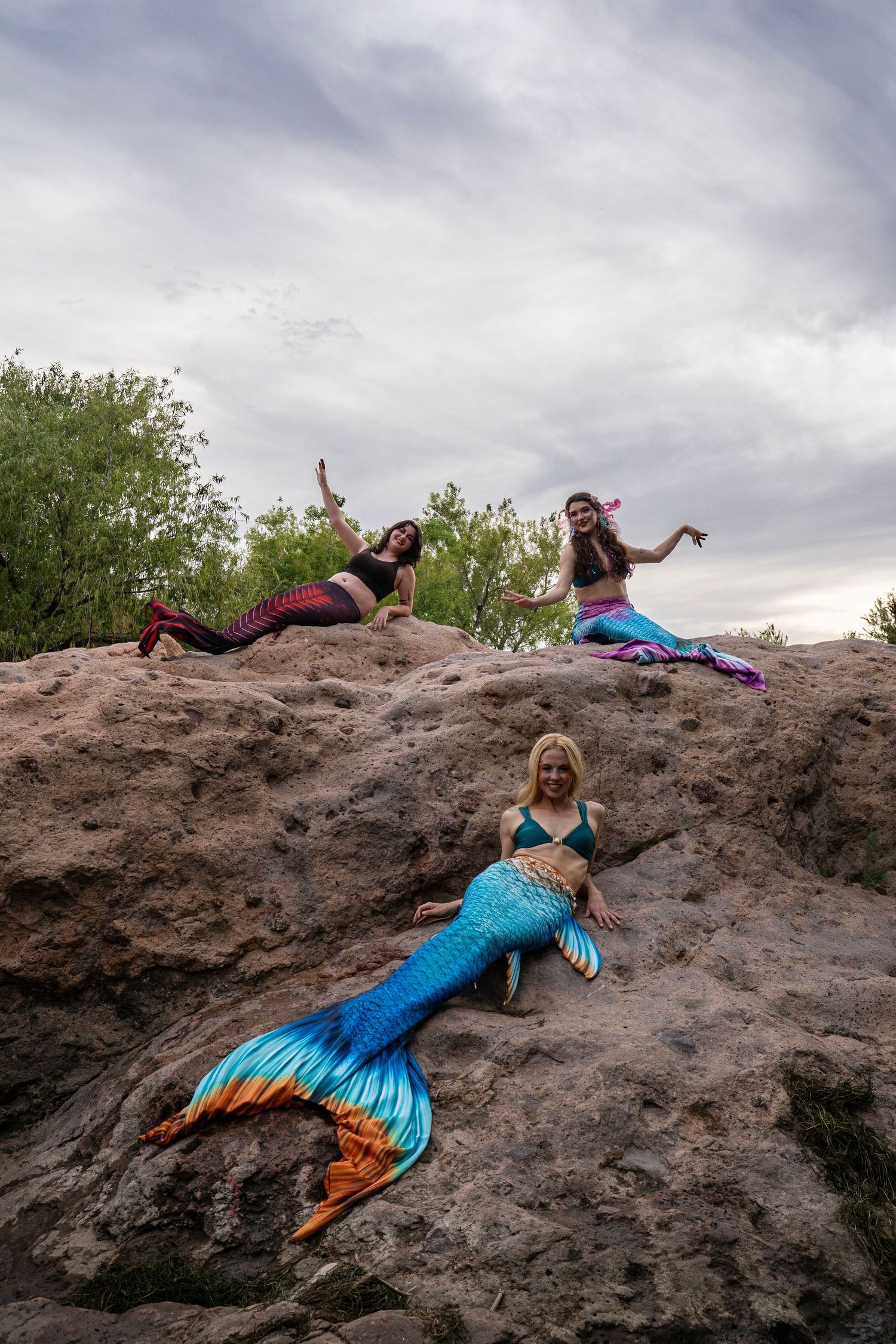 Three women in colorful mermaid costumes strike poses on large rocks against a cloudy sky. Fantasy Photoshoot