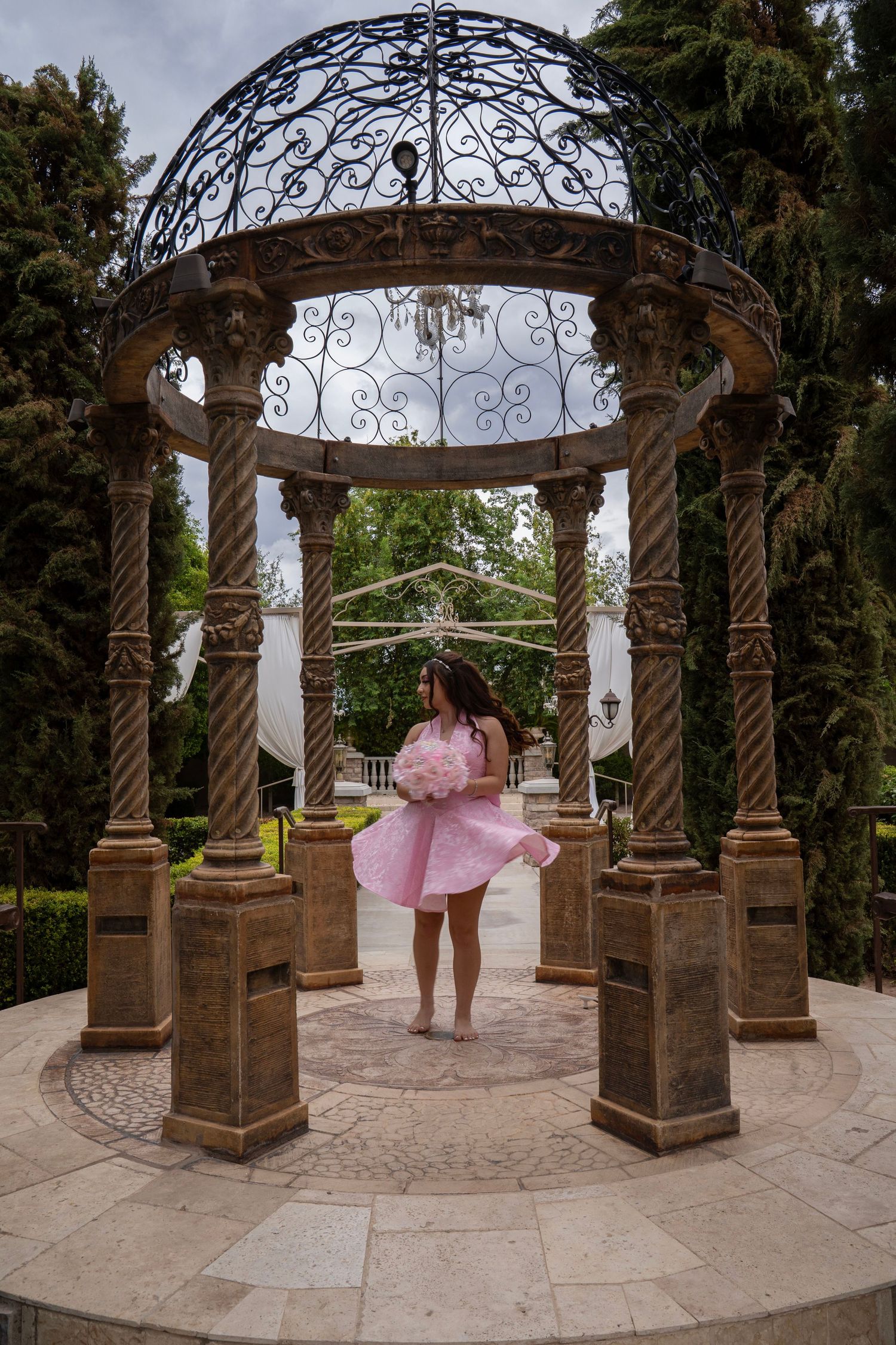 Young woman in pink dress twirling inside ornate iron gazebo with chandelier in a lush garden.