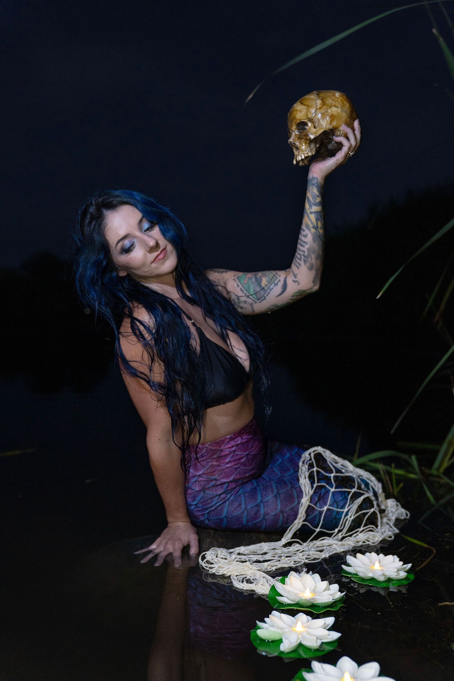 Tattooed woman in mermaid costume holds skull beside water lilies at night.