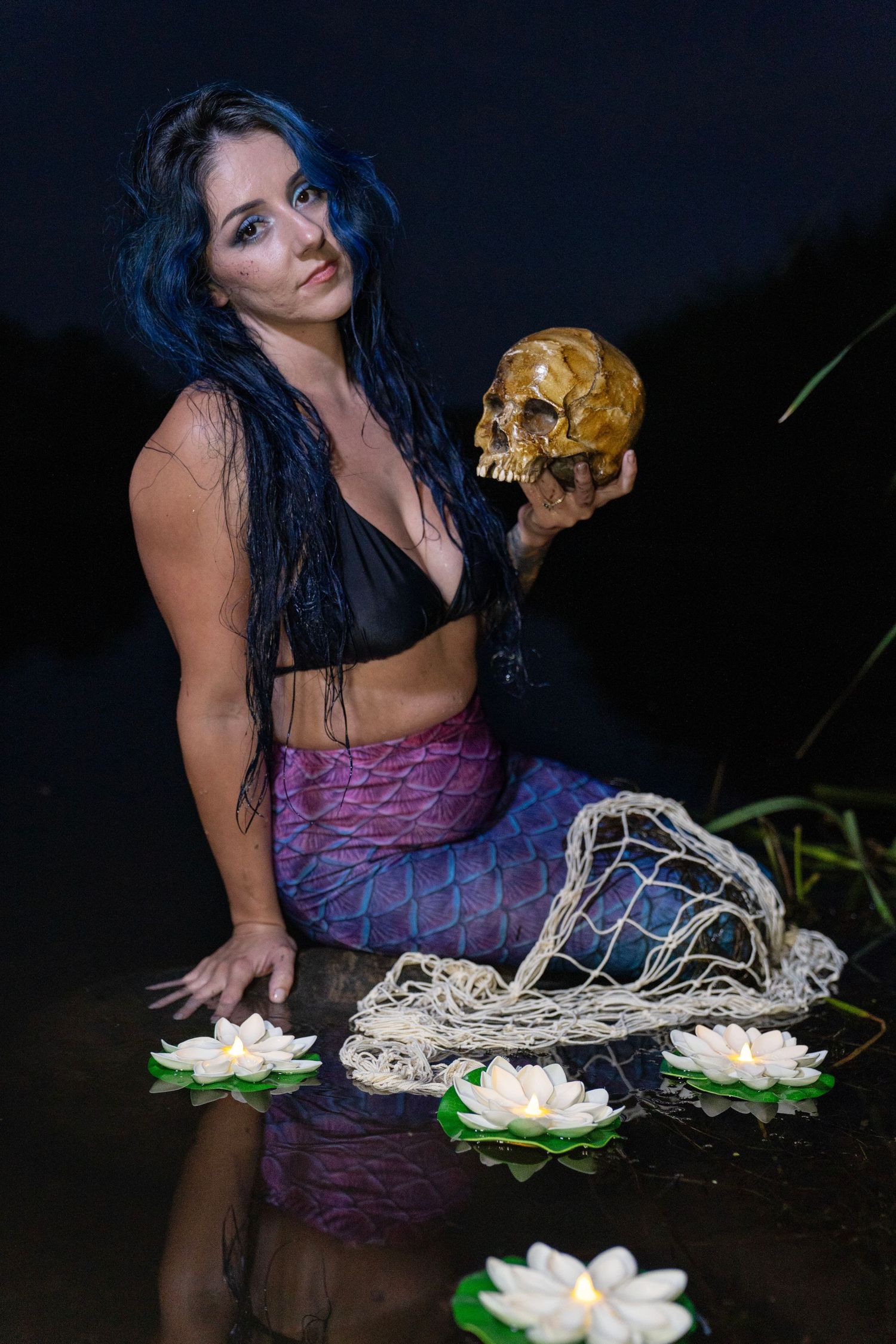 Woman in a purple mermaid tail and black top sits in dark water holding a golden skull, surrounded by floating lotus flowers and netting.