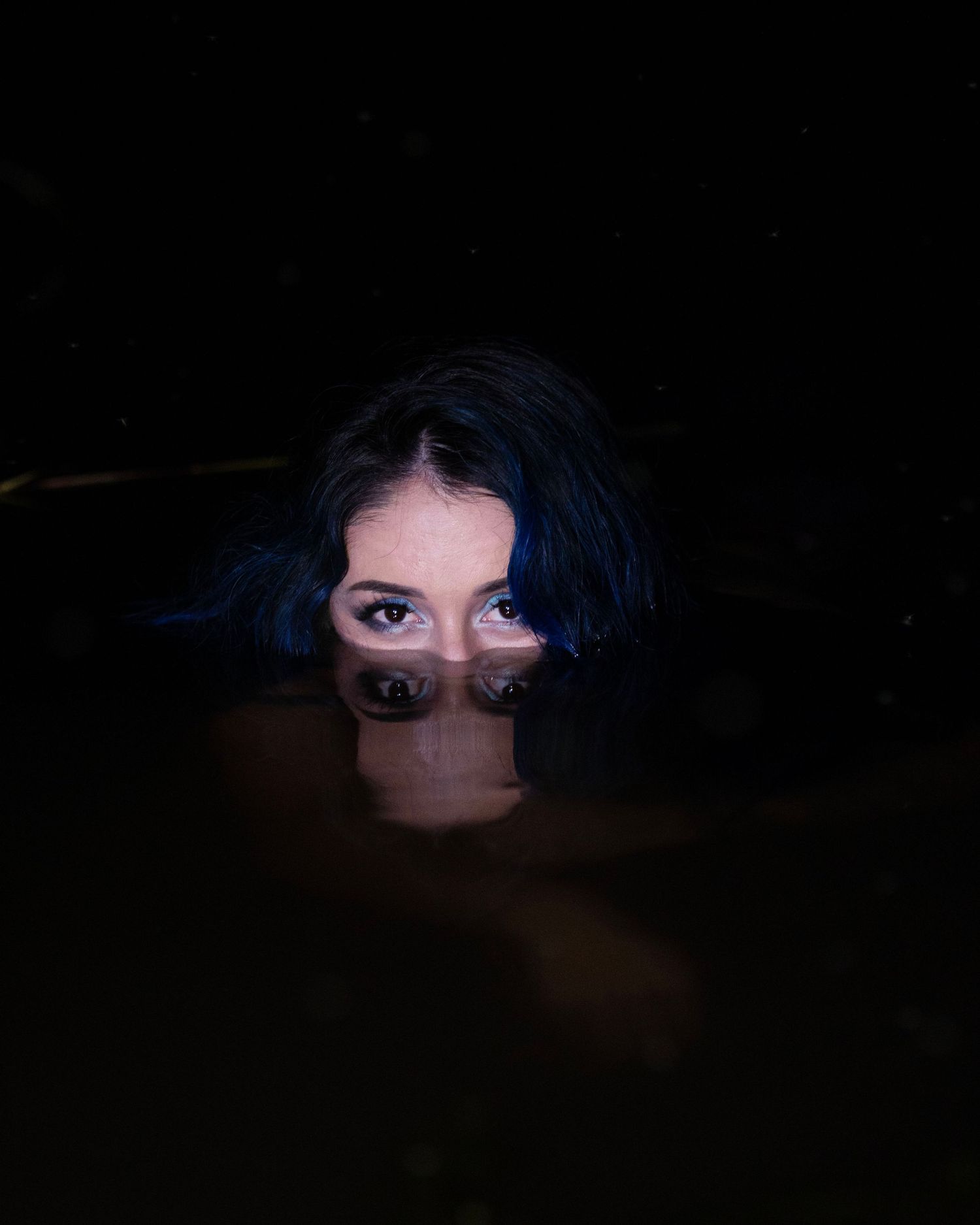 Woman with dark blue hair partially submerged in still dark water, her eyes and forehead reflected on the surface.
