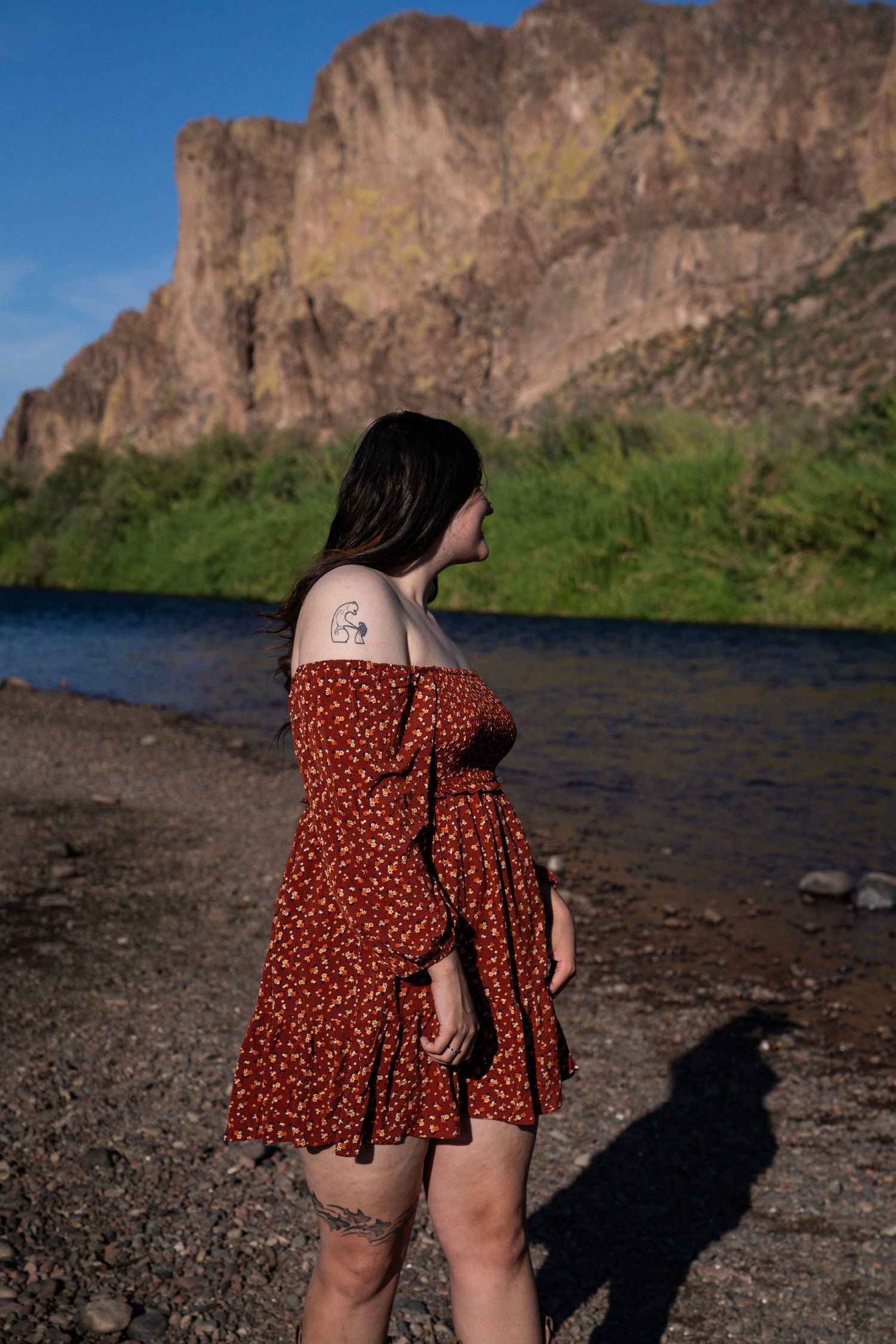 Woman in red floral off-shoulder dress standing by a river with rocky cliffs in the background.