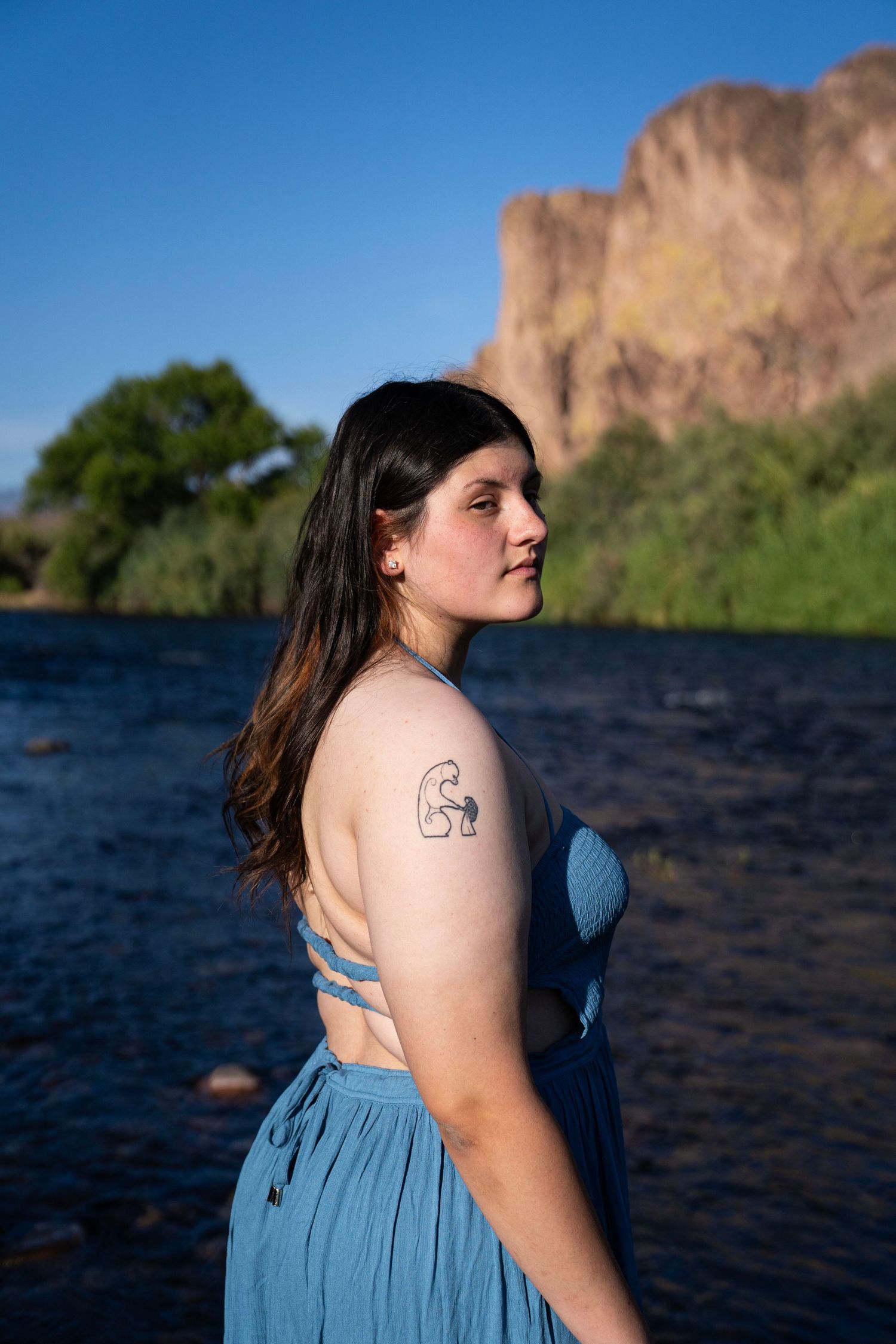 Young woman in blue backless dress with tattoo stands by river with rocky cliffs and trees in background.