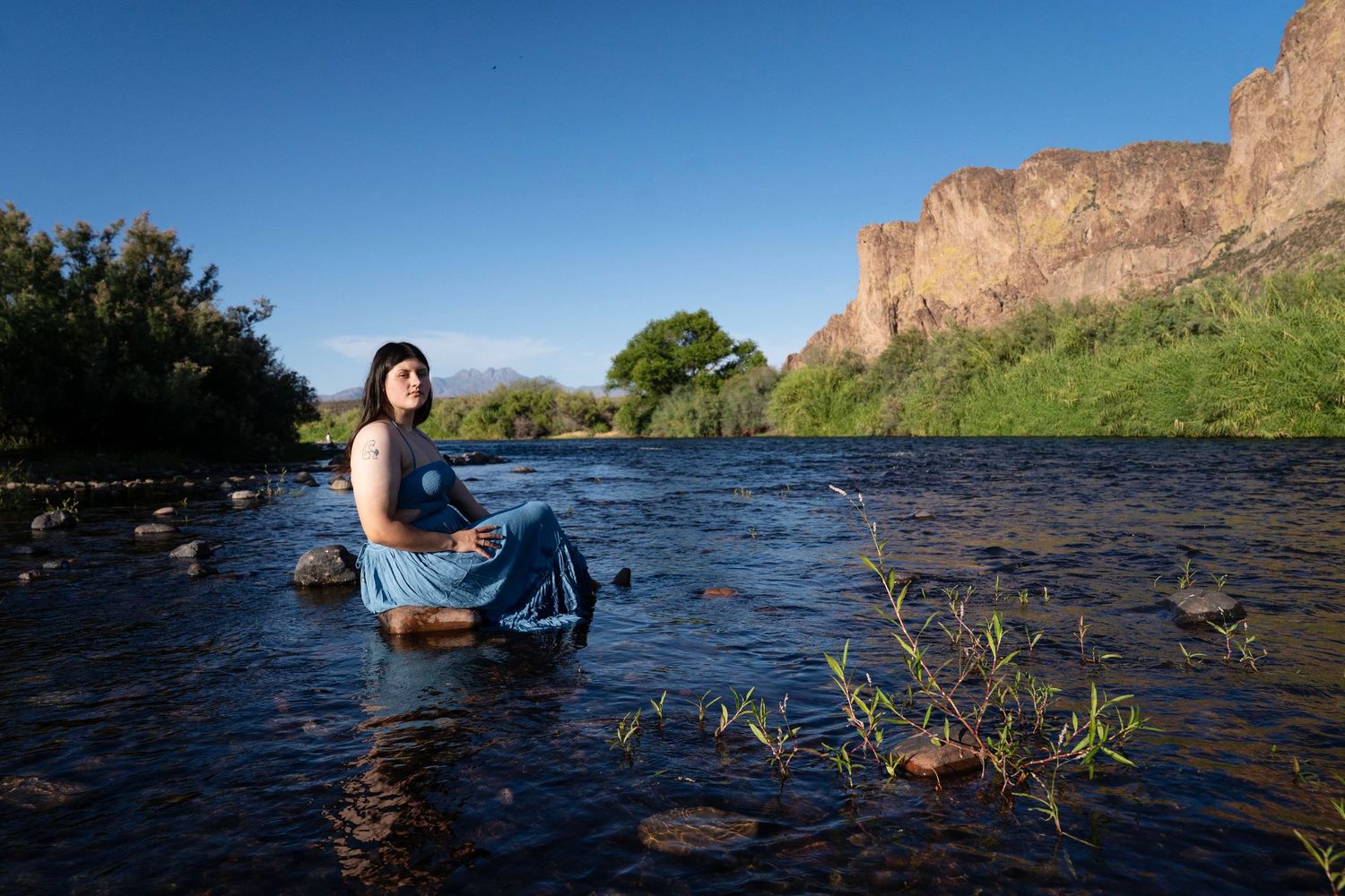 Woman in blue dress sitting on rocks in a flowing river with dramatic red cliffs and blue sky backdrop.