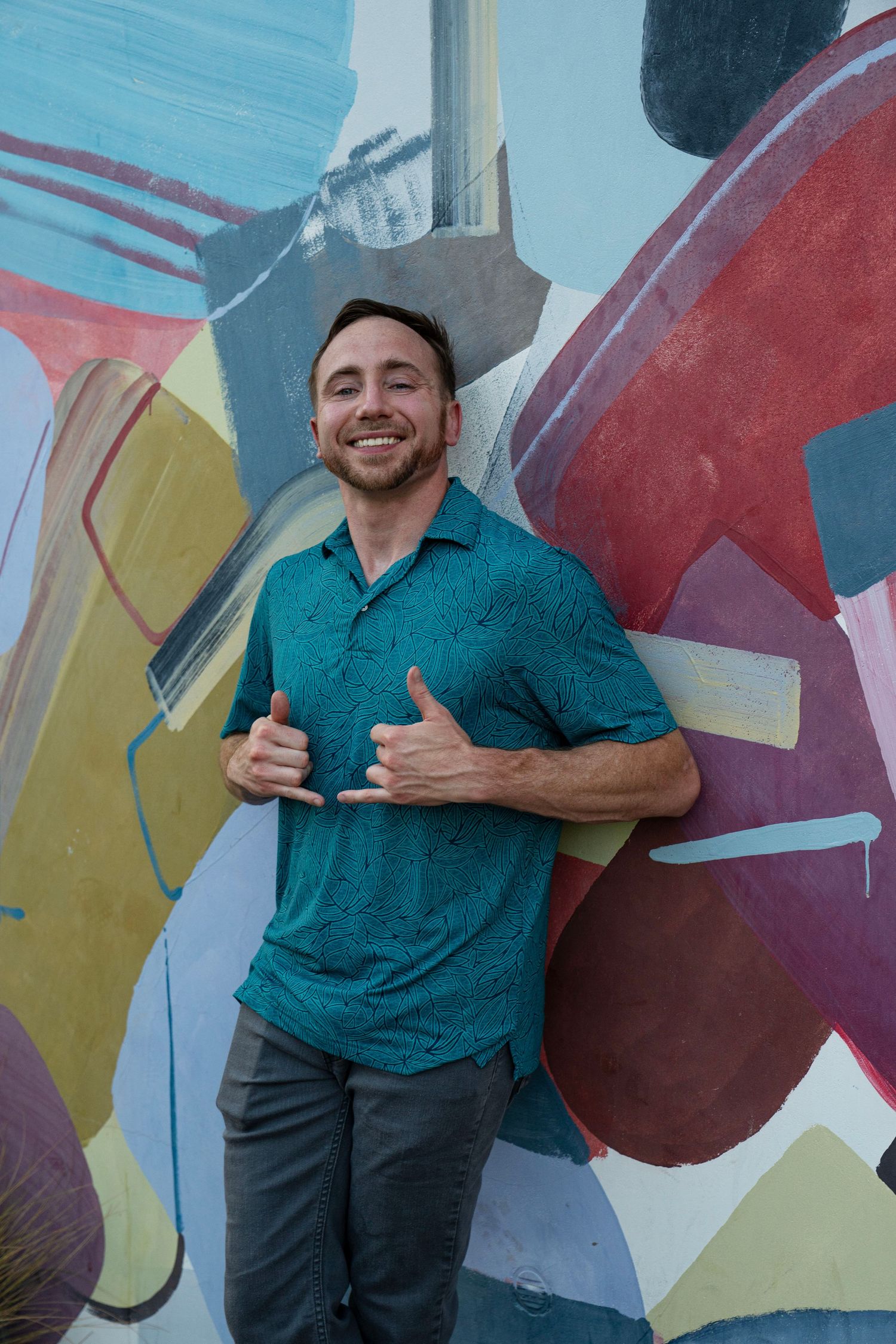 Smiling man in teal polo shirt giving thumbs up in front of colorful abstract mural wall art.
