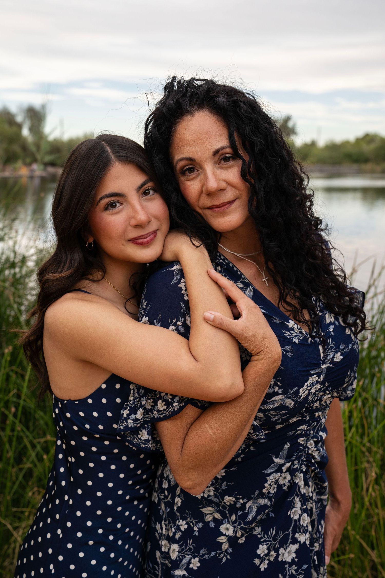 Two women in matching blue dresses embrace warmly near a lakeside, smiling for a portrait outdoors.