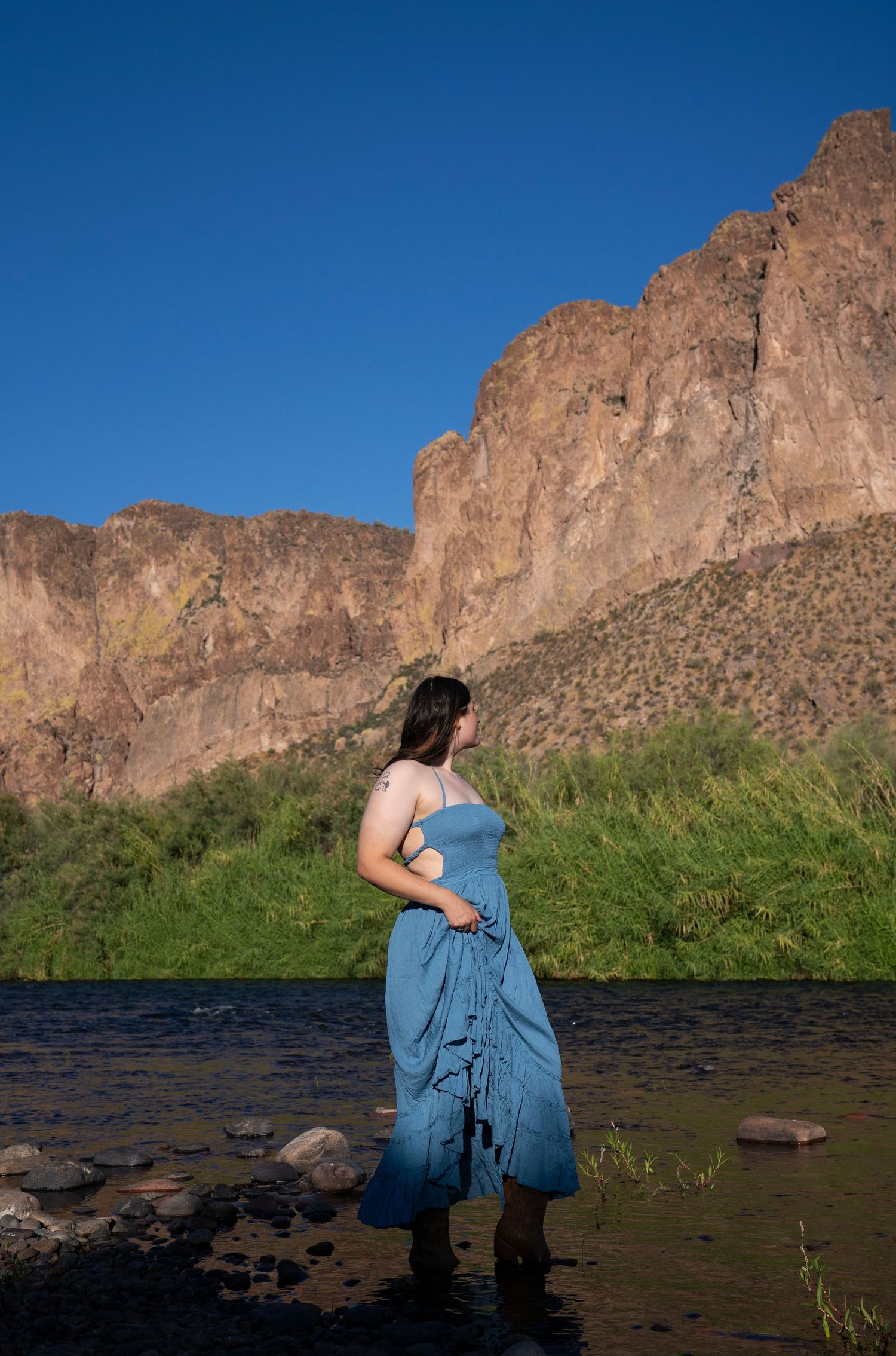 Woman in blue dress standing in a river with rocky mountains and green grass under a clear blue sky.