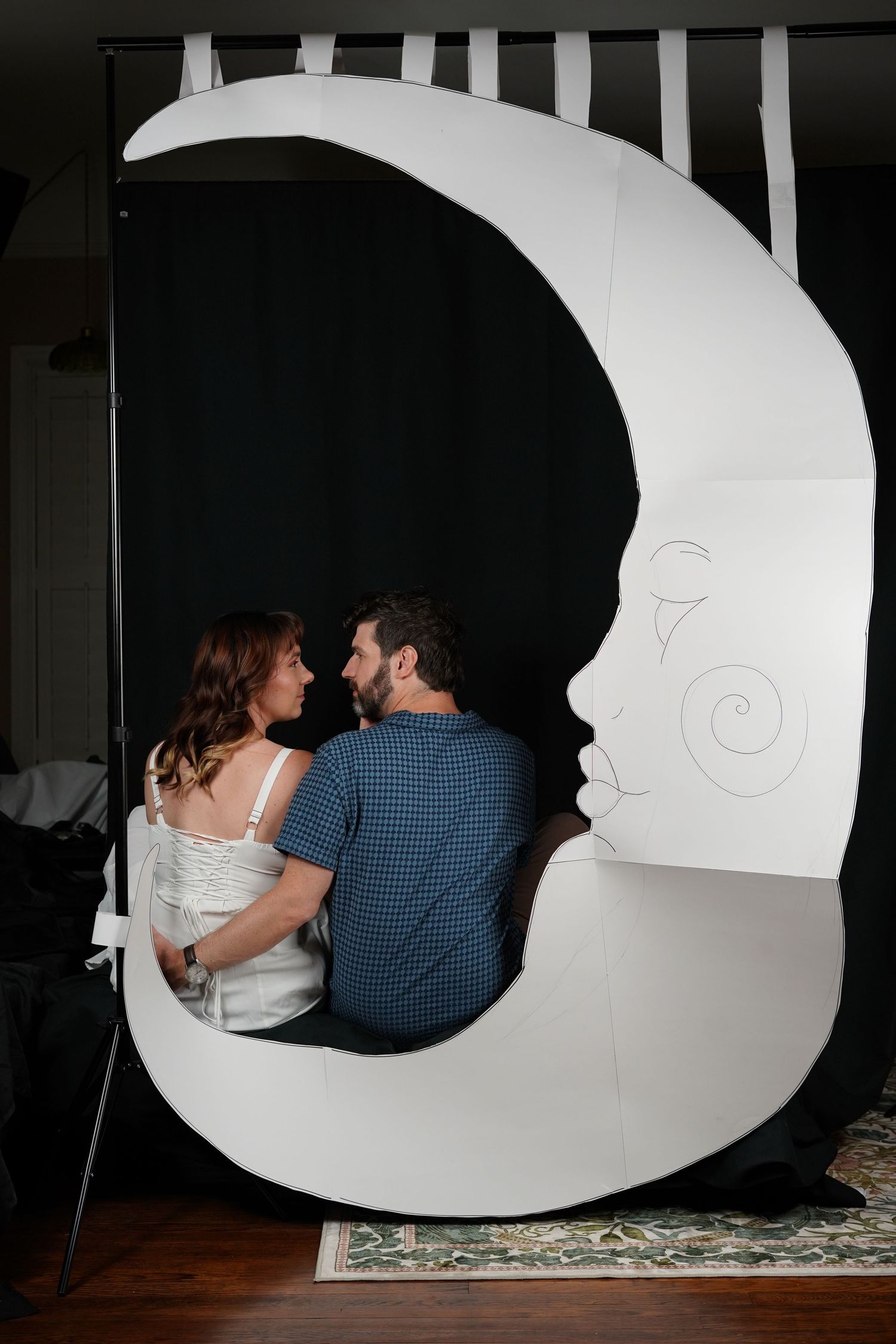 Couple sitting together in a large crescent moon prop with a face drawn on it, against a black backdrop.