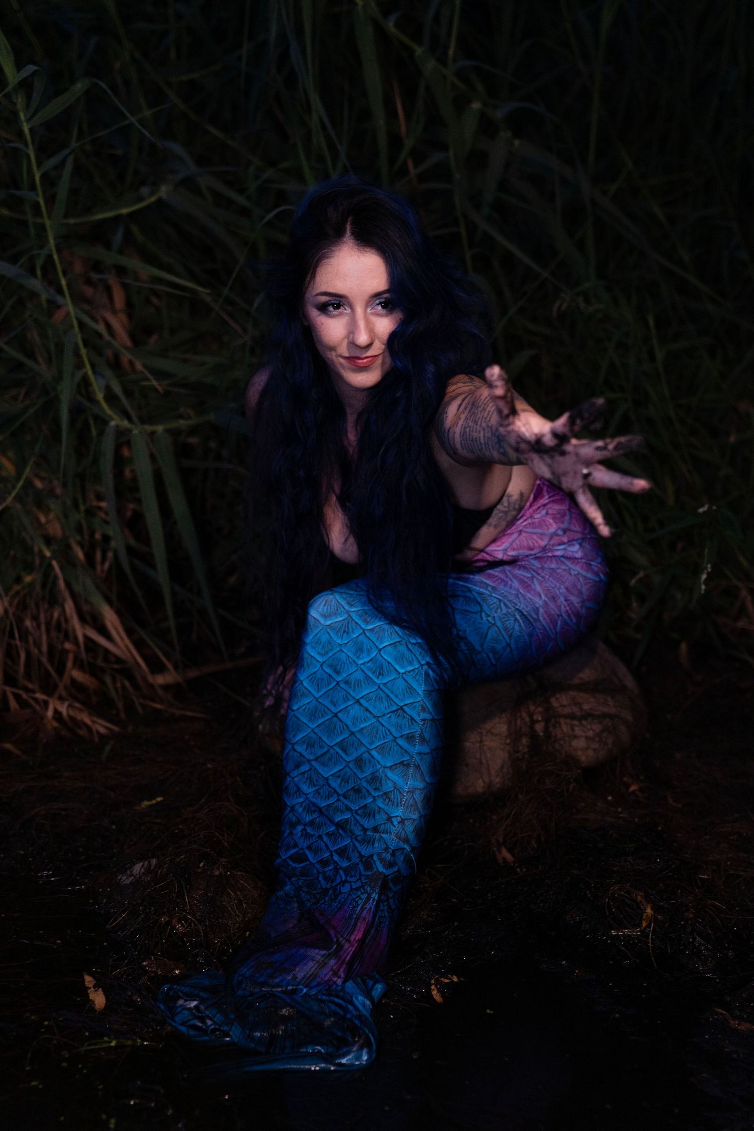 Tattooed woman in blue-pink mermaid tail crouches on rock reaching toward camera in dark setting.